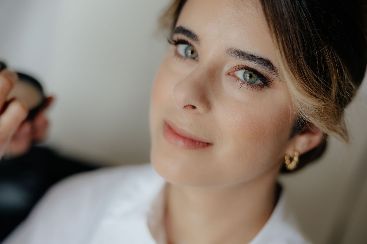 Beautiful bride with green eyes and gold earrings getting makeup done before her wedding ceremony.
