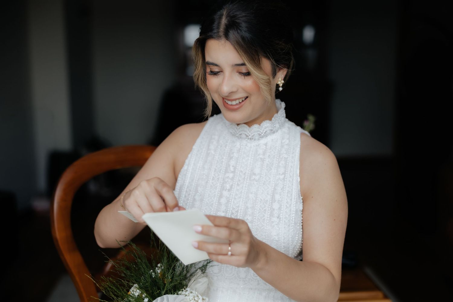 Joyful bride in a white lace gown smiling while opening an envelope with a bouquet nearby.