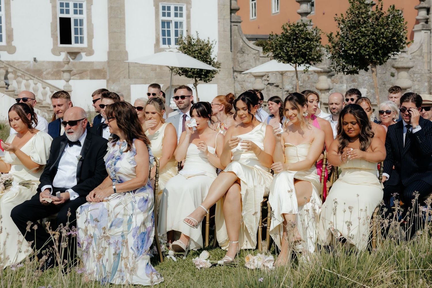 Wedding party sits together in a grassy field on a sunny day for a group photo.