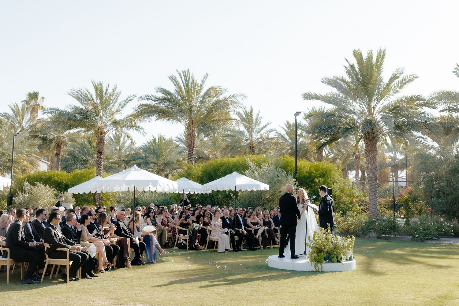 Bride and groom stand at altar on white circular platform before large crowd at outdoor palm tree wedding ceremony.