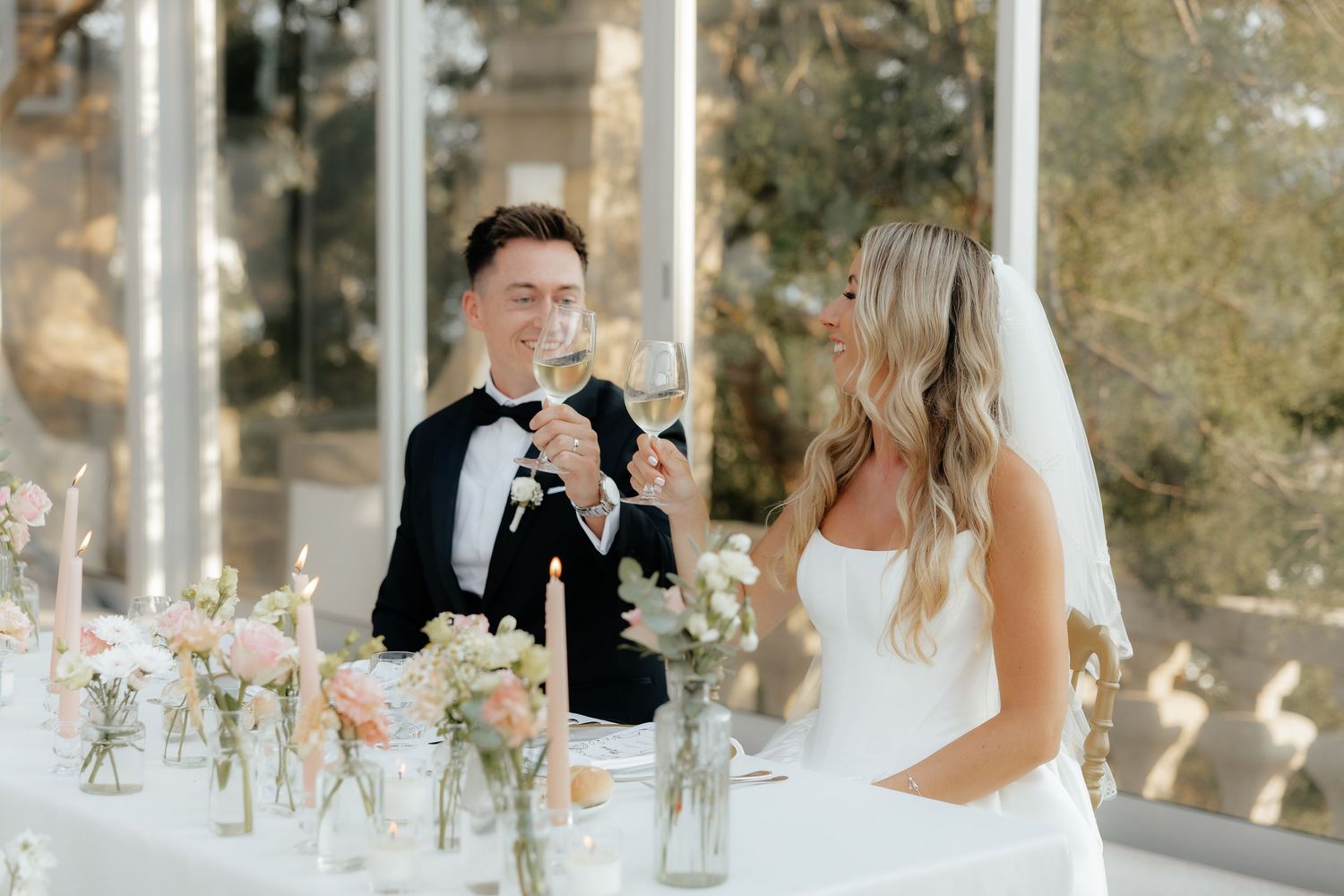 Elegant wedding reception table settings with white floral centerpieces and glowing candles in a sunlit venue.