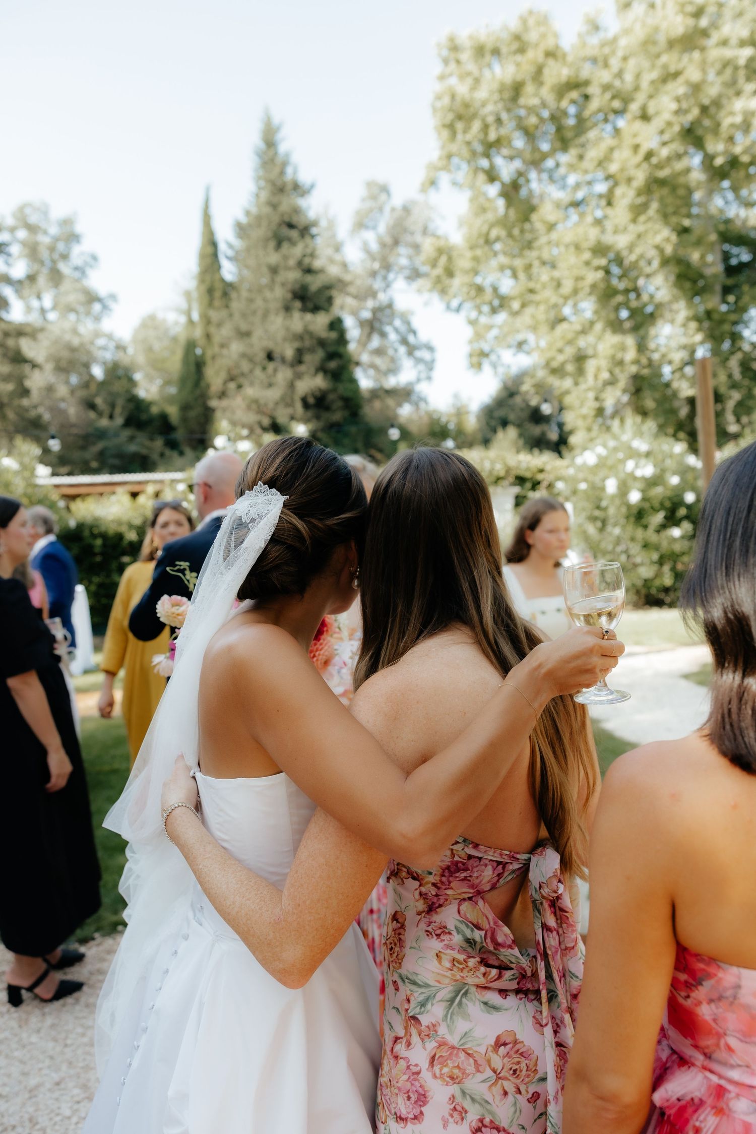 Wedding guests gather outdoors for celebratory drinks under sunny garden trees.