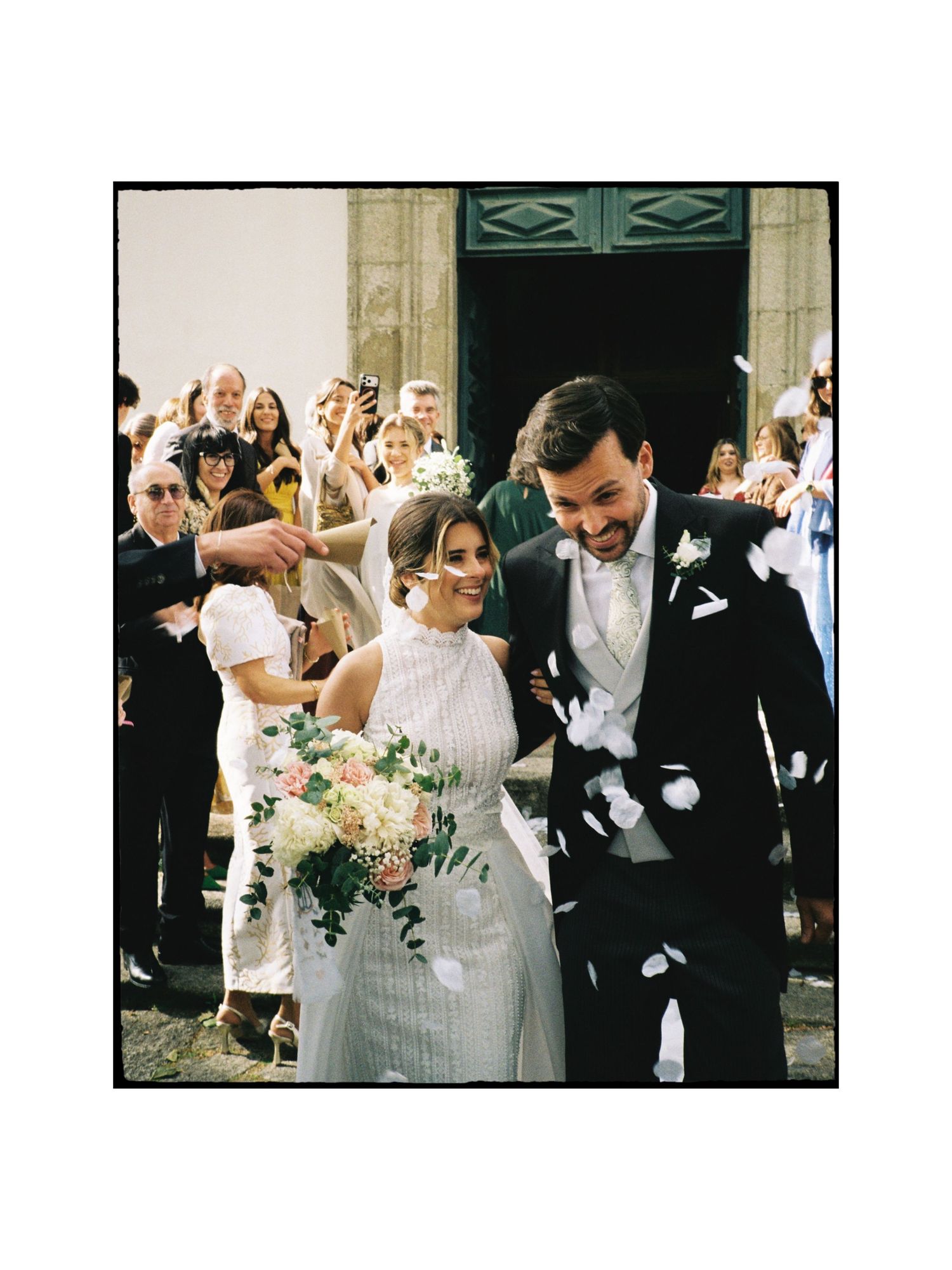 Smiling bride and groom walk through confetti shower as guests celebrate outside a church.