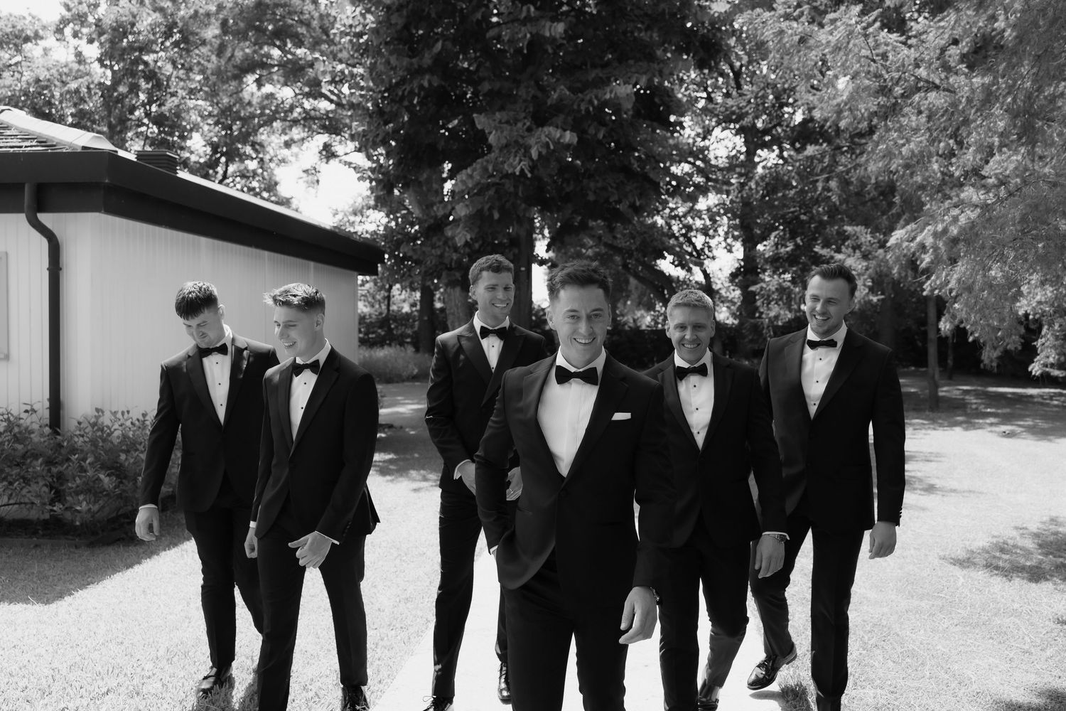 Black and white photo of groom and groomsmen in tuxedos walking together along a pathway near a white building.
