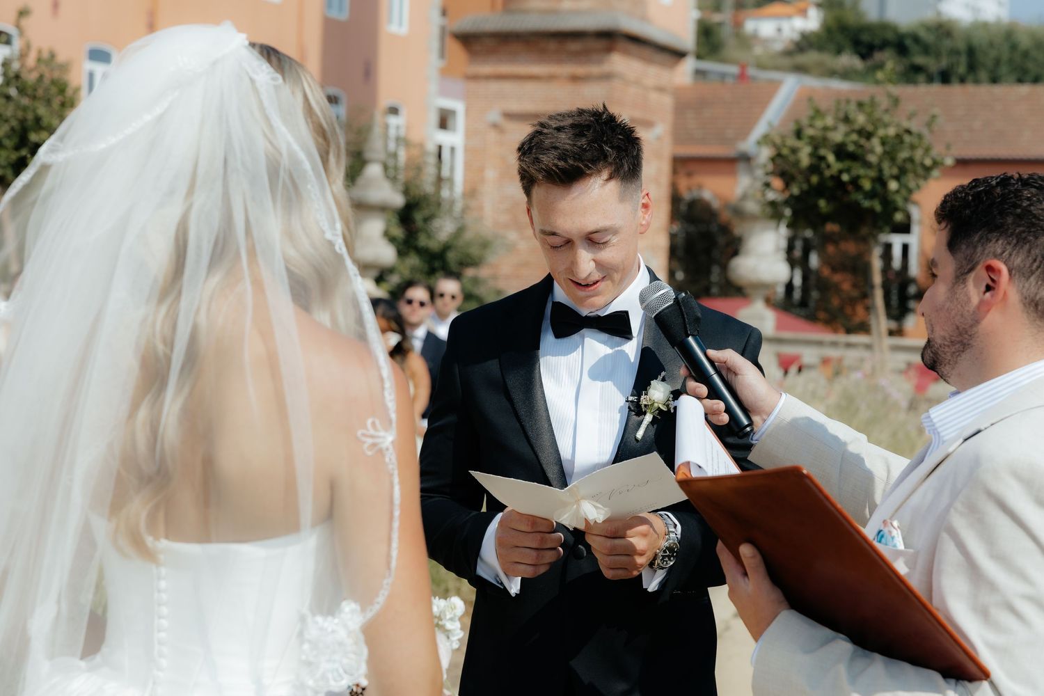 Groom reads wedding vows from paper to bride at outdoor ceremony with officiant nearby.
