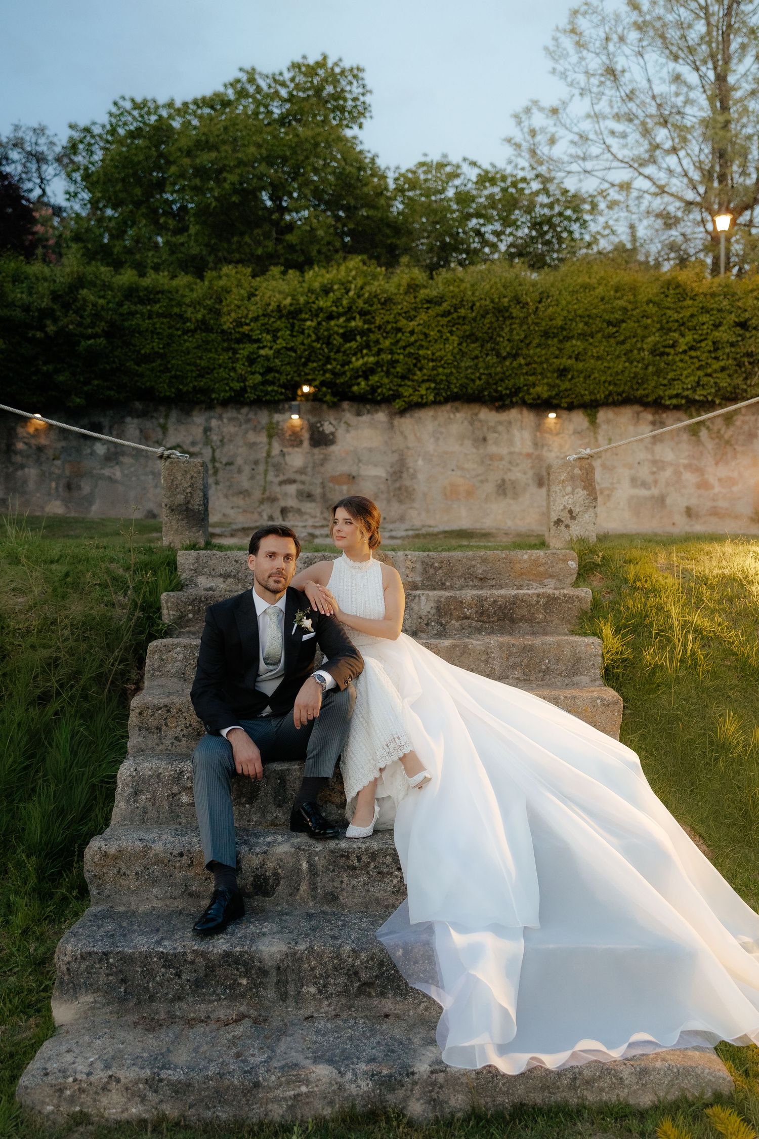 Bride and groom sitting on stone steps in garden at dusk with string lights.