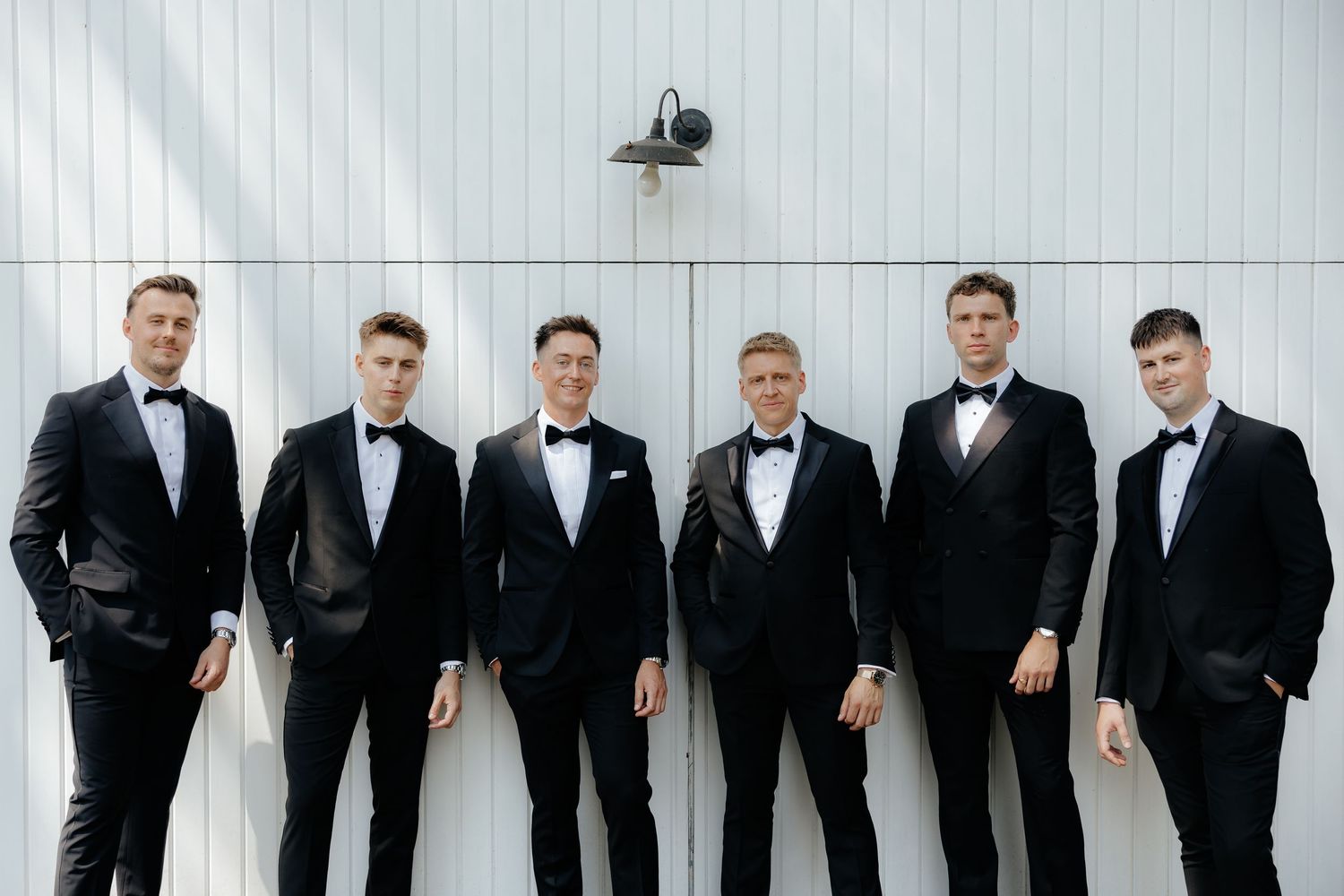 Six groomsmen in black tuxedos standing together in a relaxed pose against a white wall.