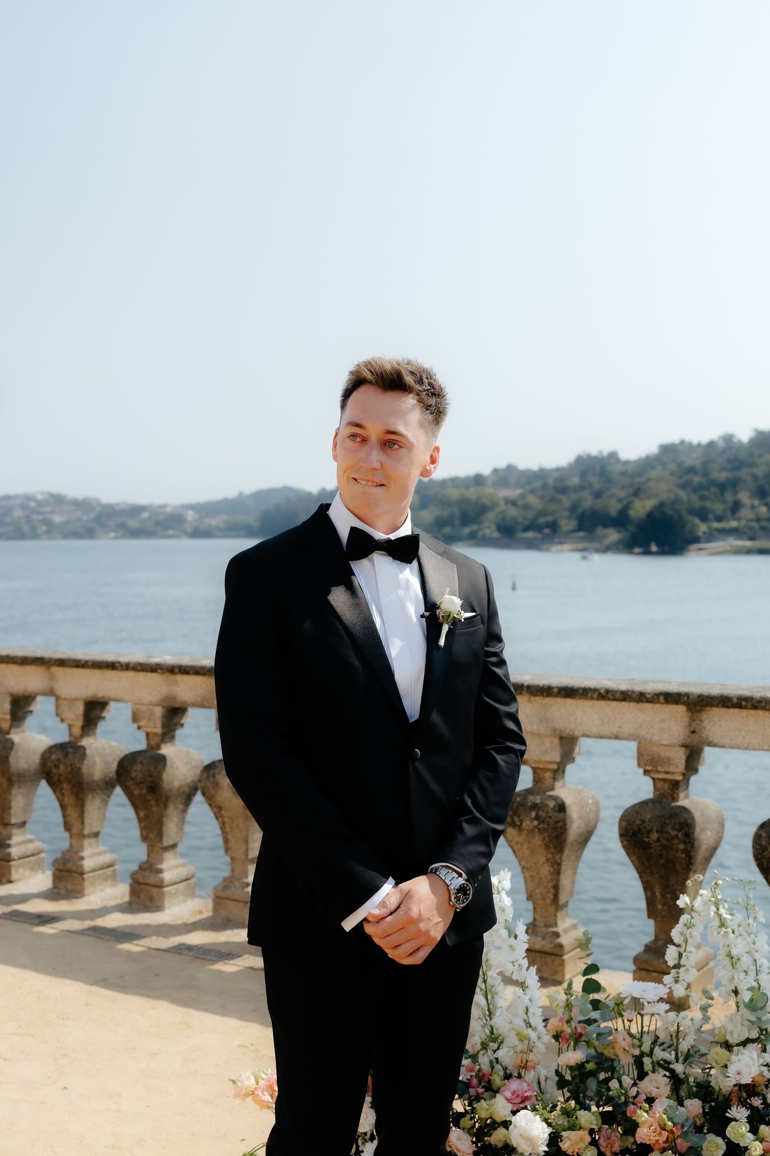 Handsome groom in black tuxedo and bow tie stands at waterfront wedding ceremony altar.