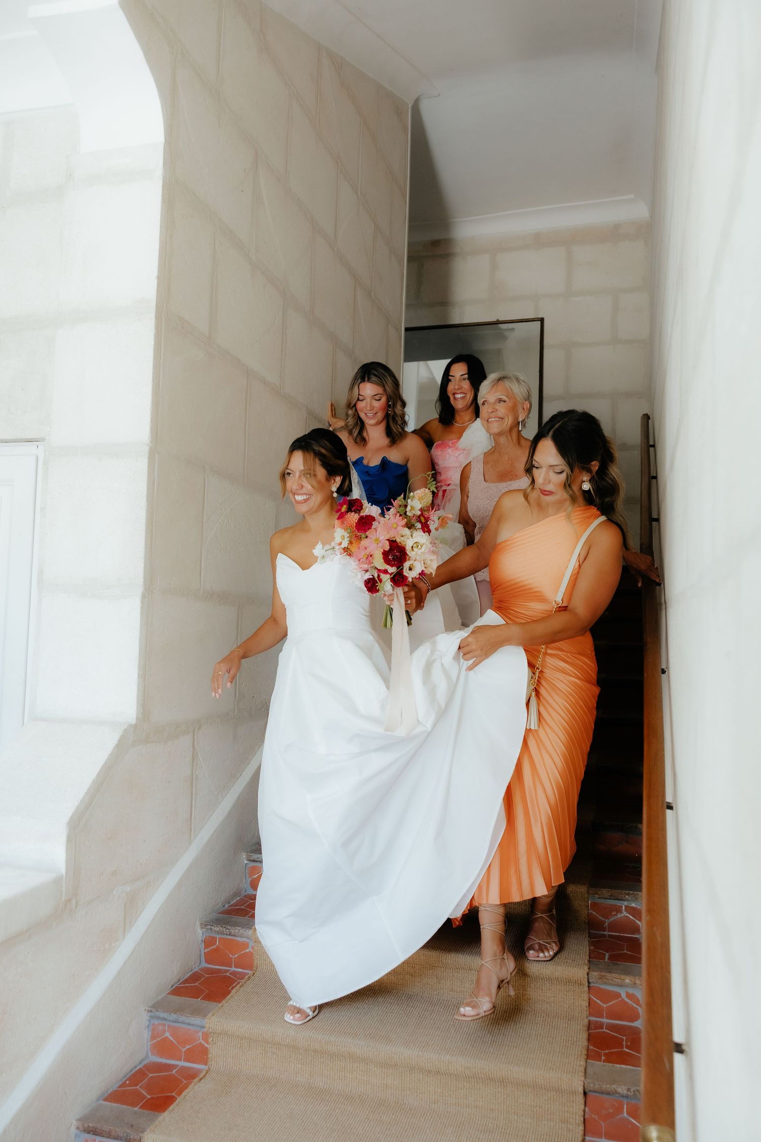 A wedding party ascending stairs in orange and pink dresses while helping with the bride's gown.