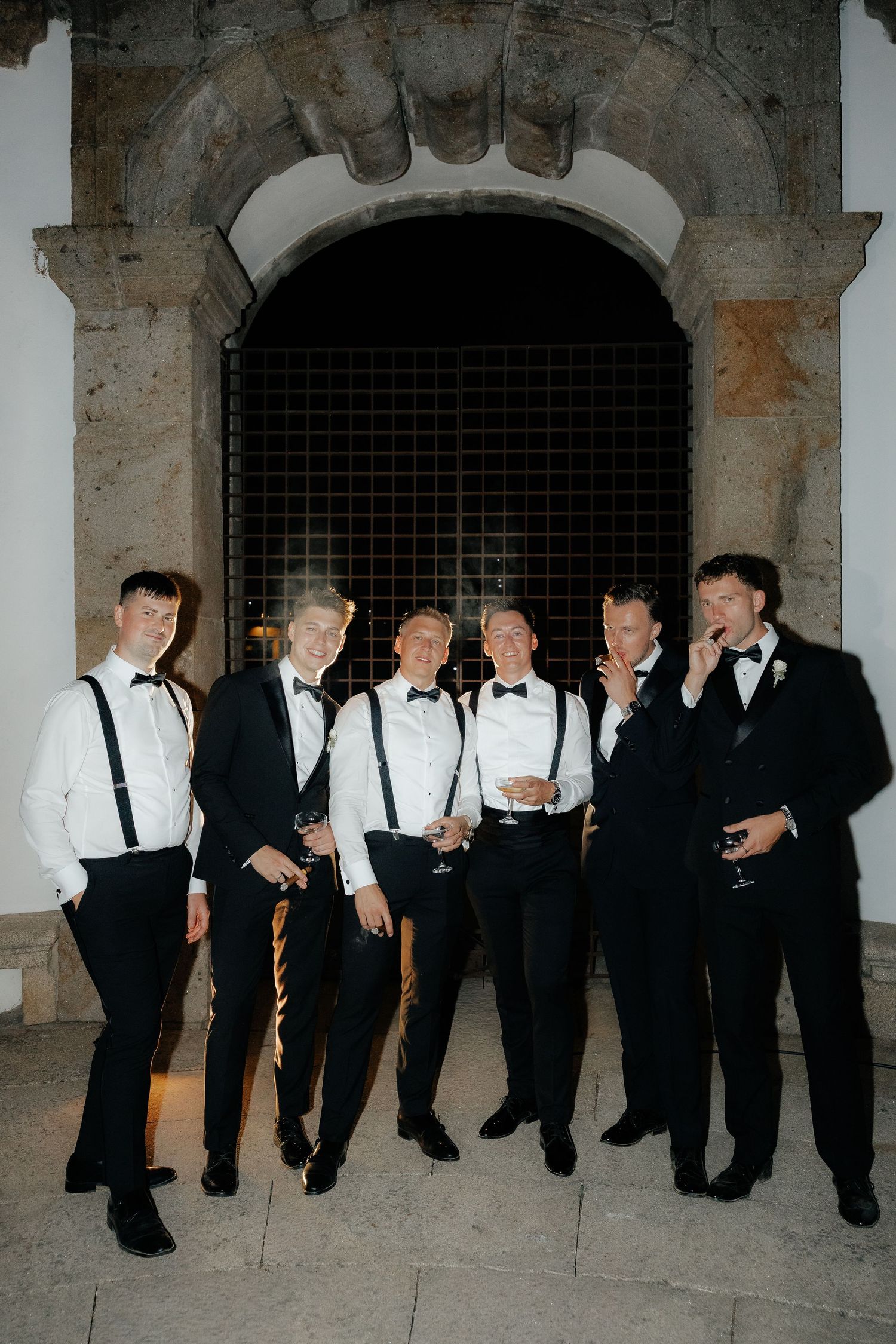 Groomsmen in black suits and white suspenders pose together in a rustic stone archway at a wedding reception.