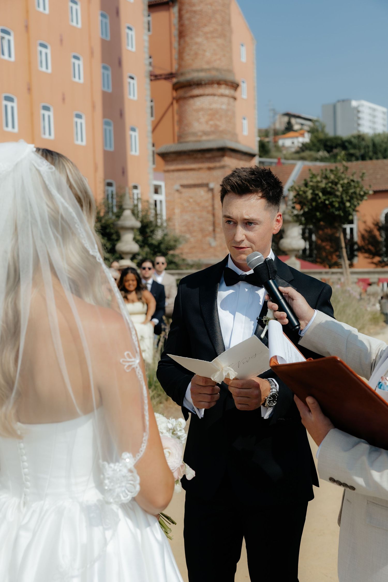 A couple in formal wedding attire stands together outdoors near a brick building during their ceremony.