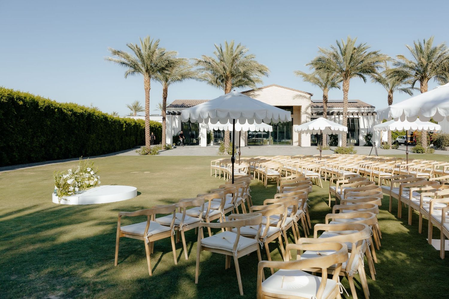 Outdoor wedding ceremony setup with wooden chairs, white umbrellas, palm trees, and a white pavilion backdrop on green lawn.