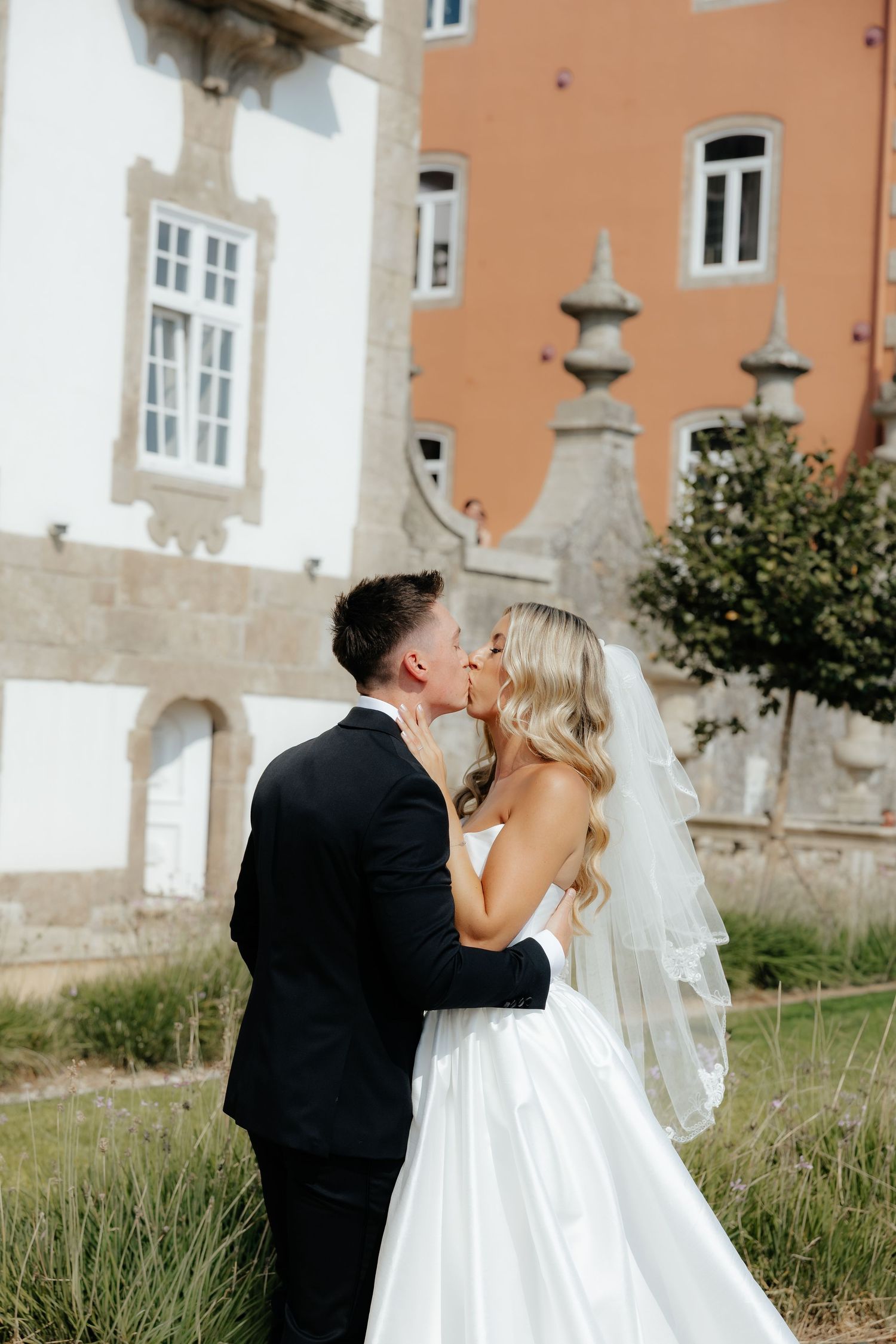 A romantic wedding couple shares an intimate kiss against a rustic stone building backdrop.