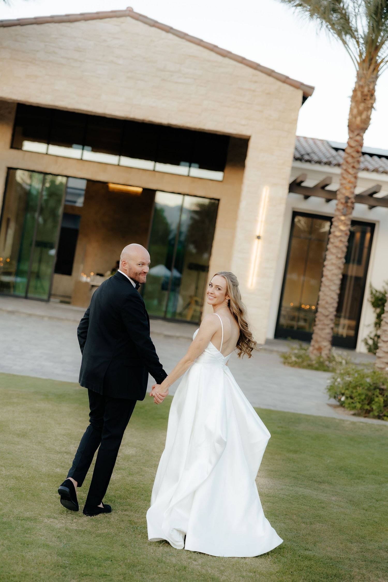 Bride in white gown and groom in tuxedo holding hands walking across lawn, smiling back at camera.