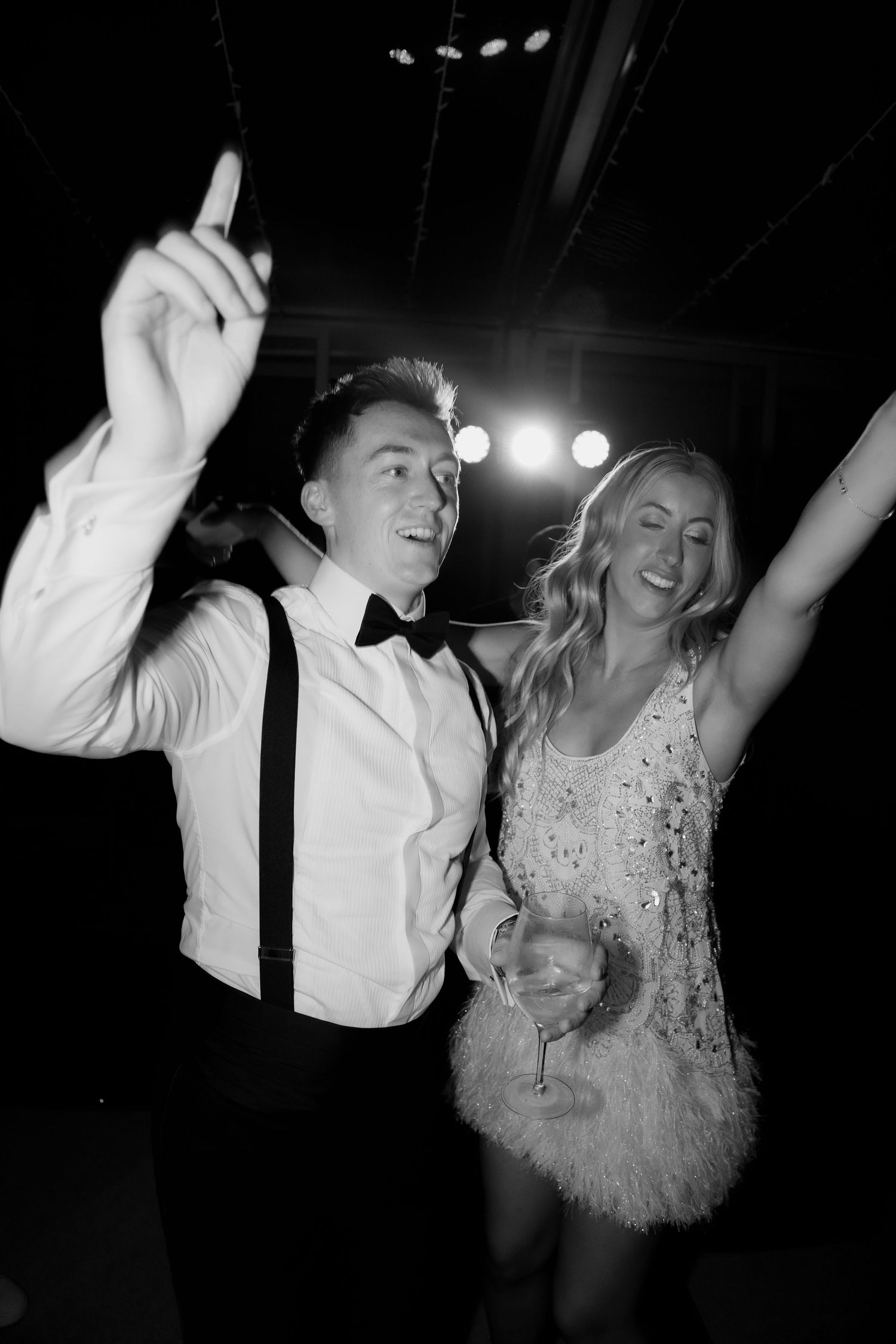 A couple dances joyfully at a wedding reception in black and white, with the bride wearing a short feathered dress.
