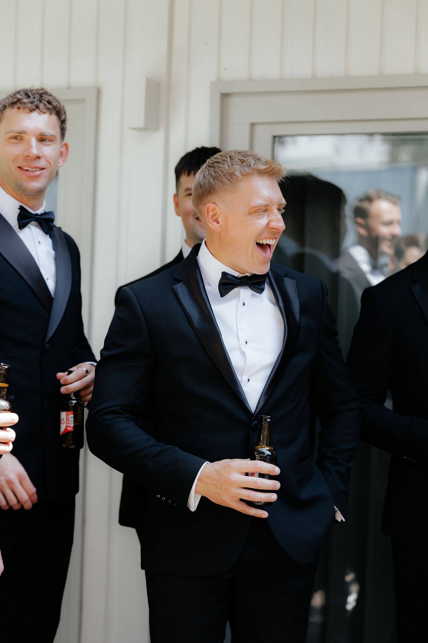 Groomsmen in black tuxedos and bowties share laughs while holding drinks at an elegant wedding celebration.