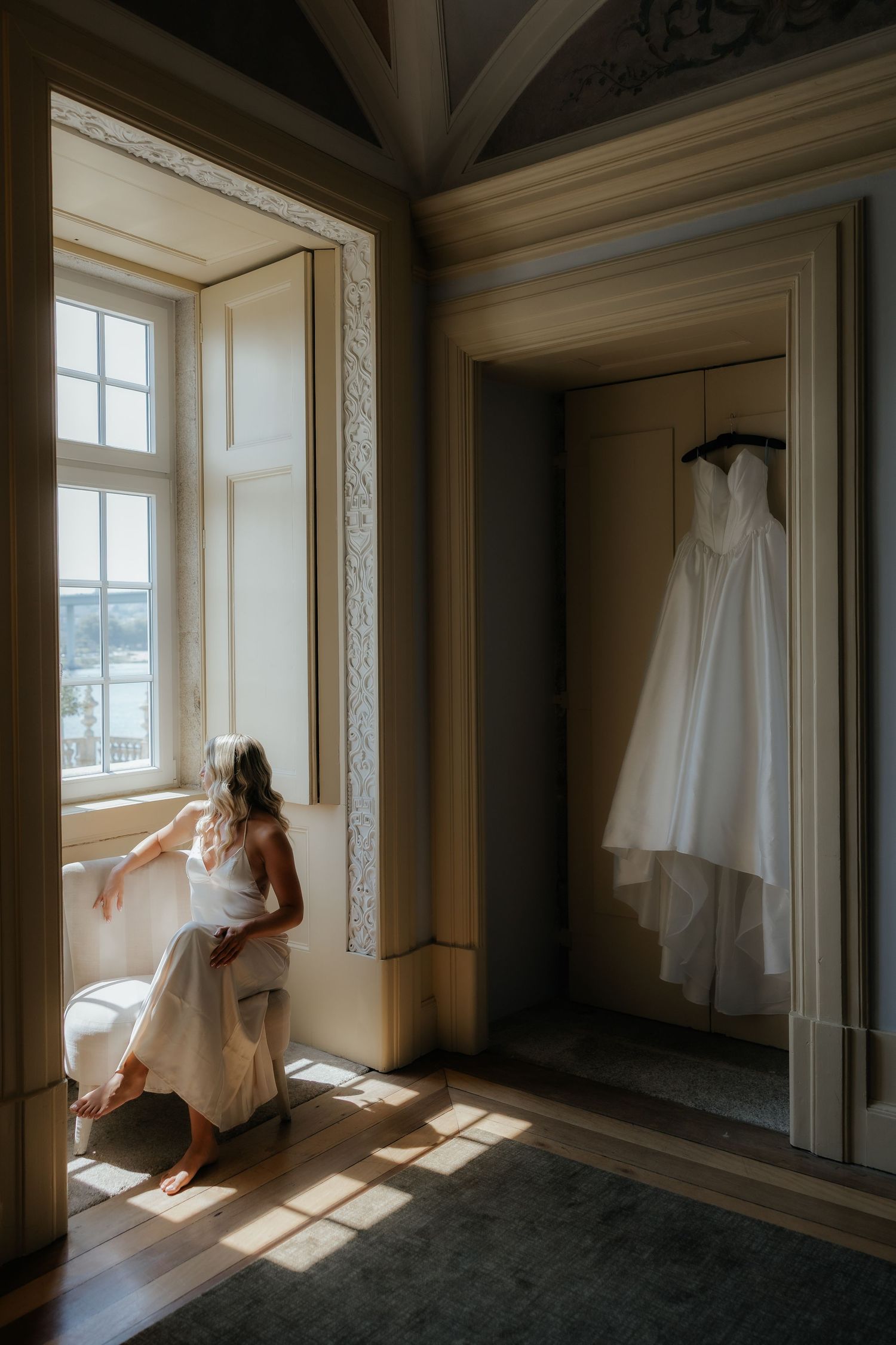 A wedding dress hangs in a doorway while sunlight streams through a window in an elegant room.