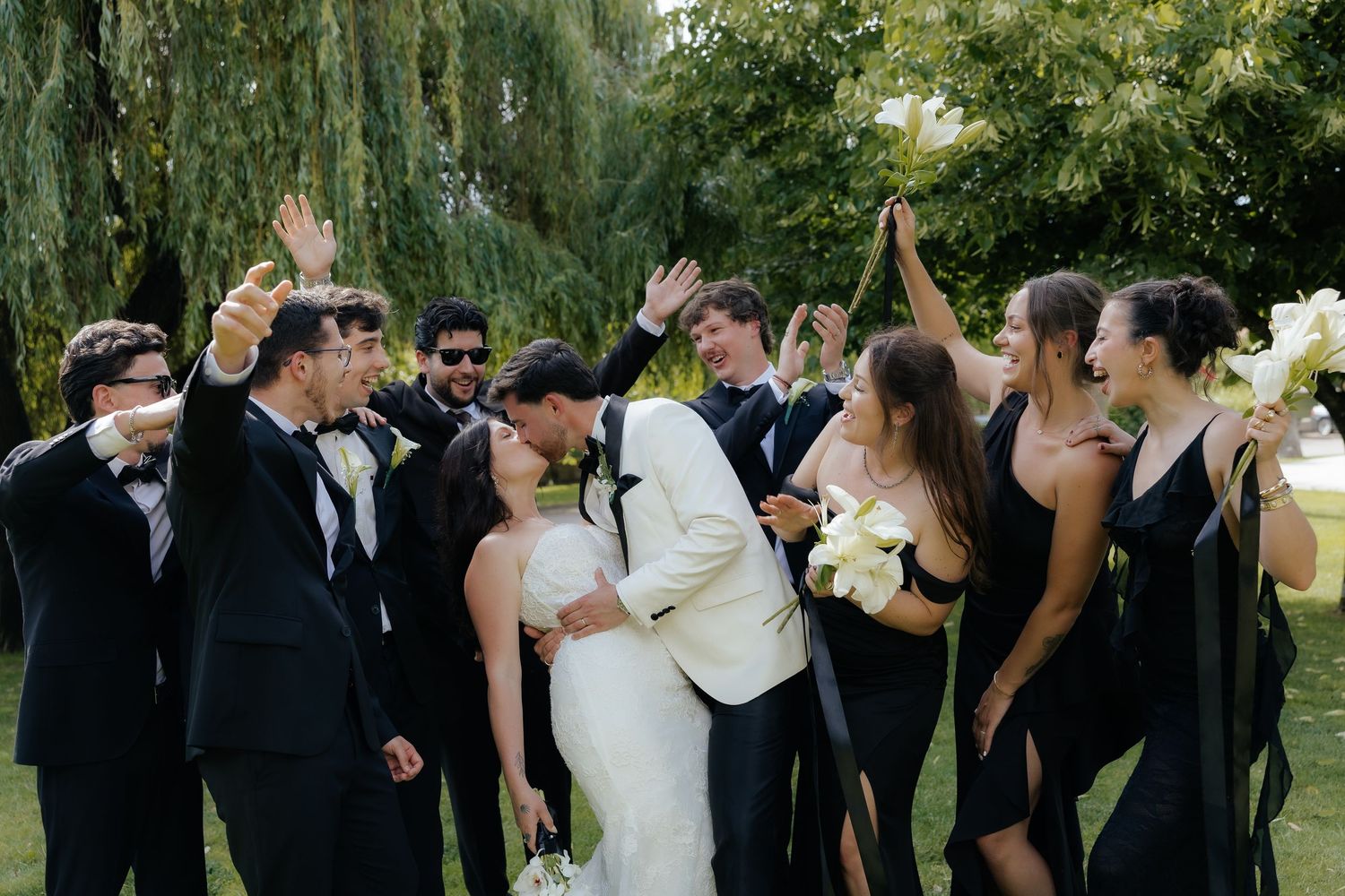 Wedding party celebrating with raised hands as bride and groom kiss on green lawn outdoors.