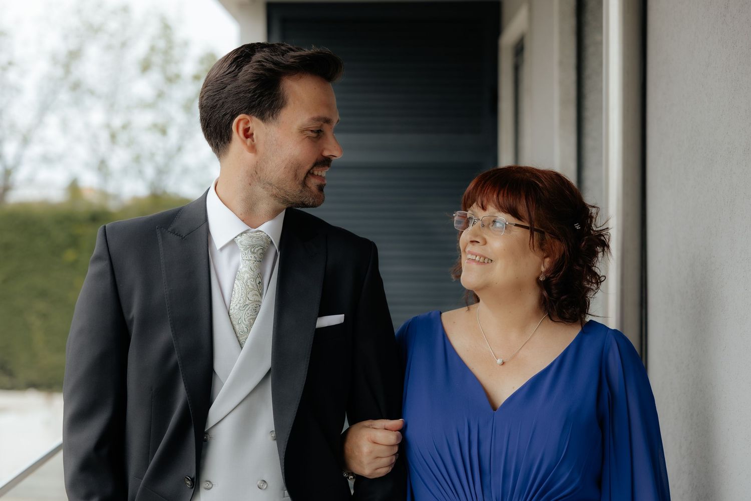 Groom in gray suit smiles at older woman in blue dress on balcony.