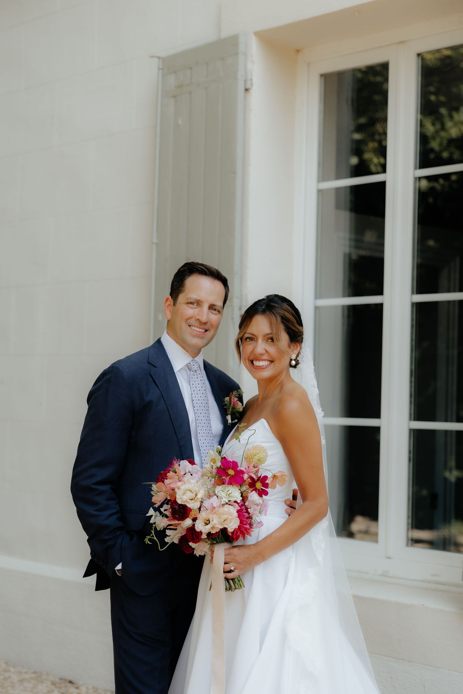 Romantic wedding portraits against white building with colorful summer bouquet.