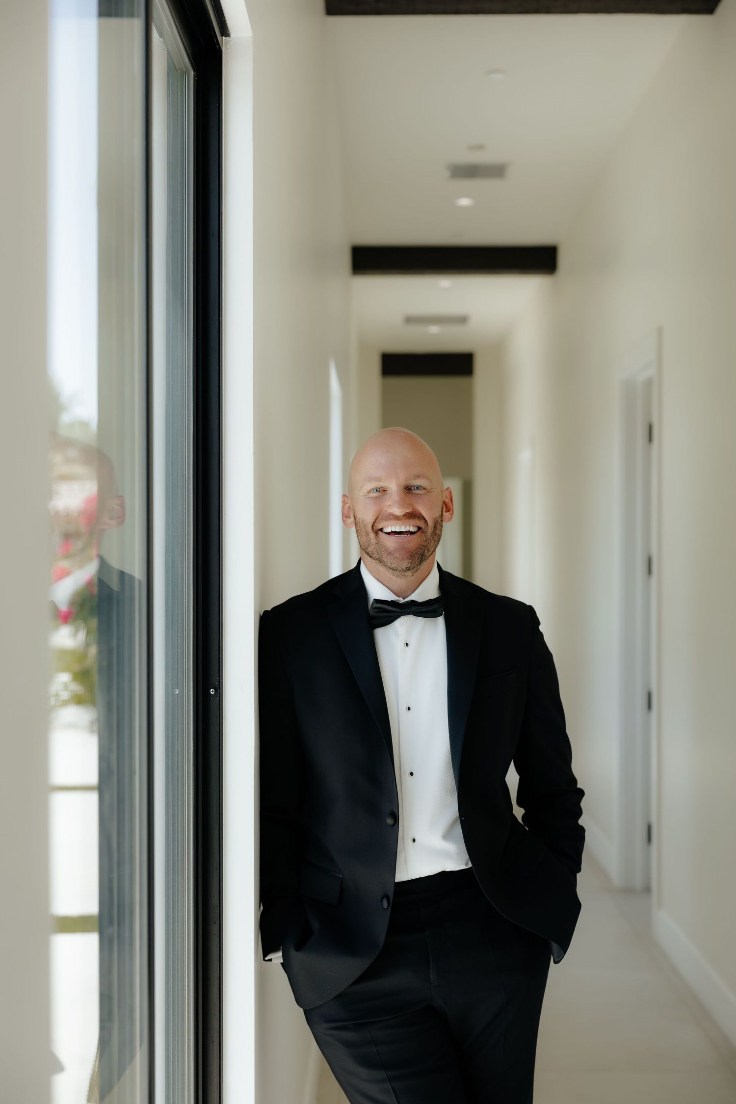 Laughing groom in black tuxedo and bow tie leans against a bright white hallway wall before his wedding.