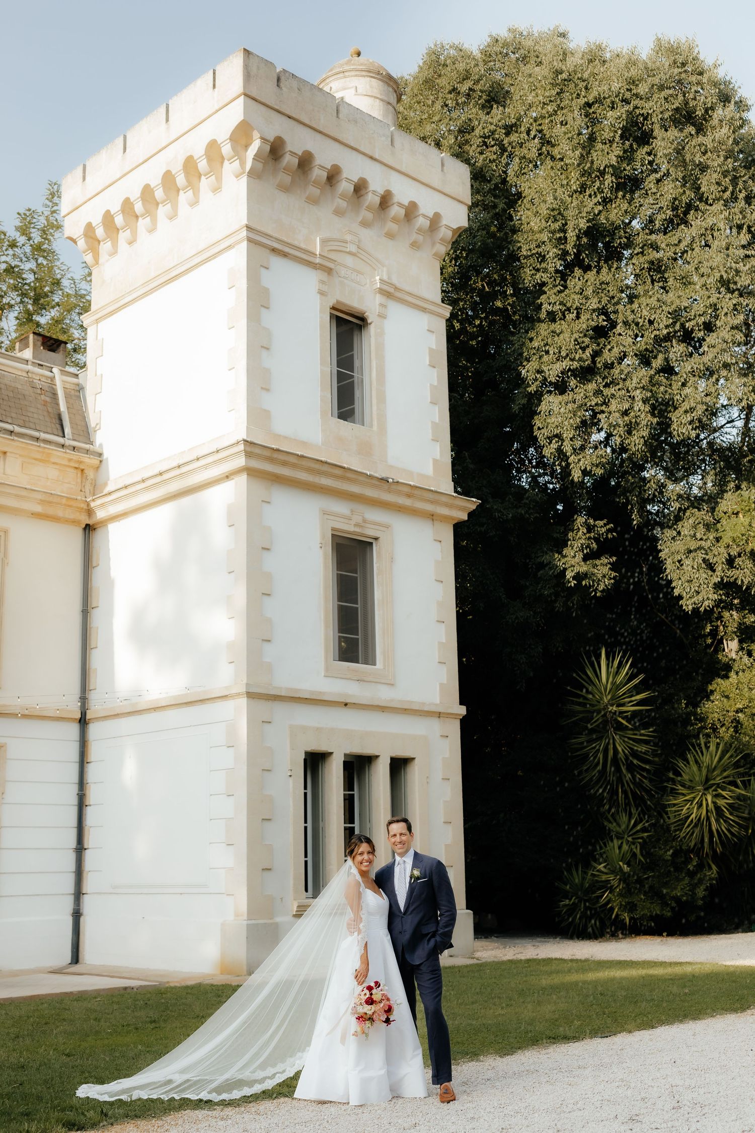 A couple poses for wedding photos in front of a white stone tower with ornate architectural details.
