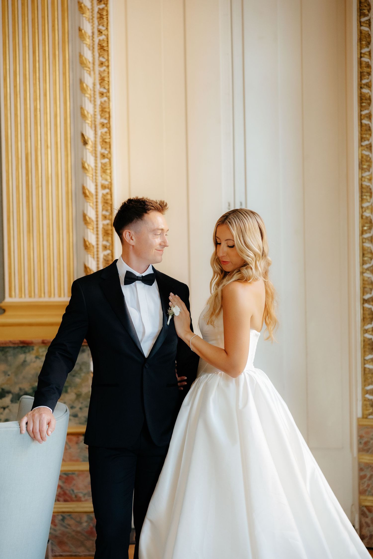 A bride in a white ballgown and groom in black tuxedo share playful moments in an elegant gold-trimmed room.