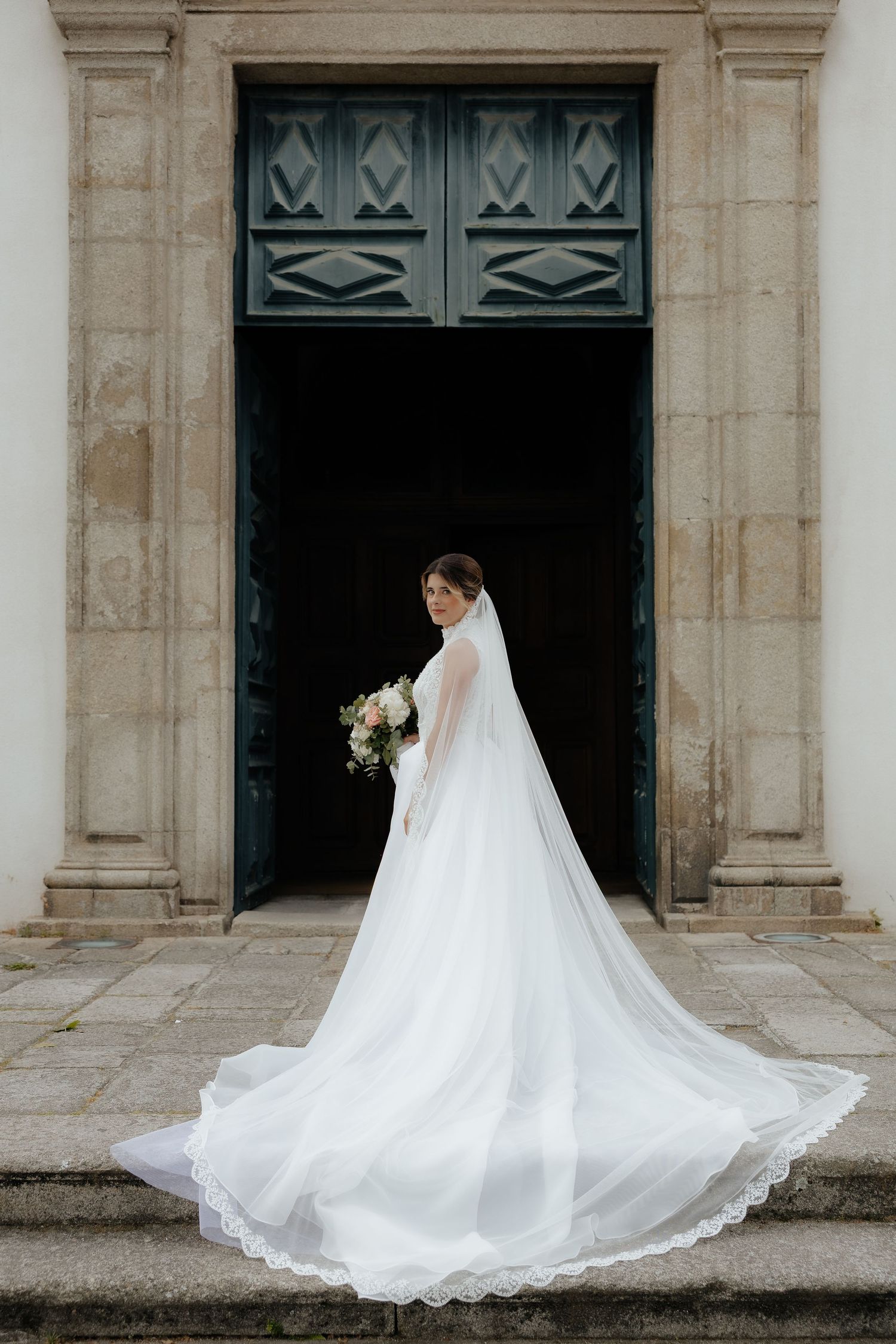 Bride in elegant white gown with long veil stands on church steps holding bouquet.