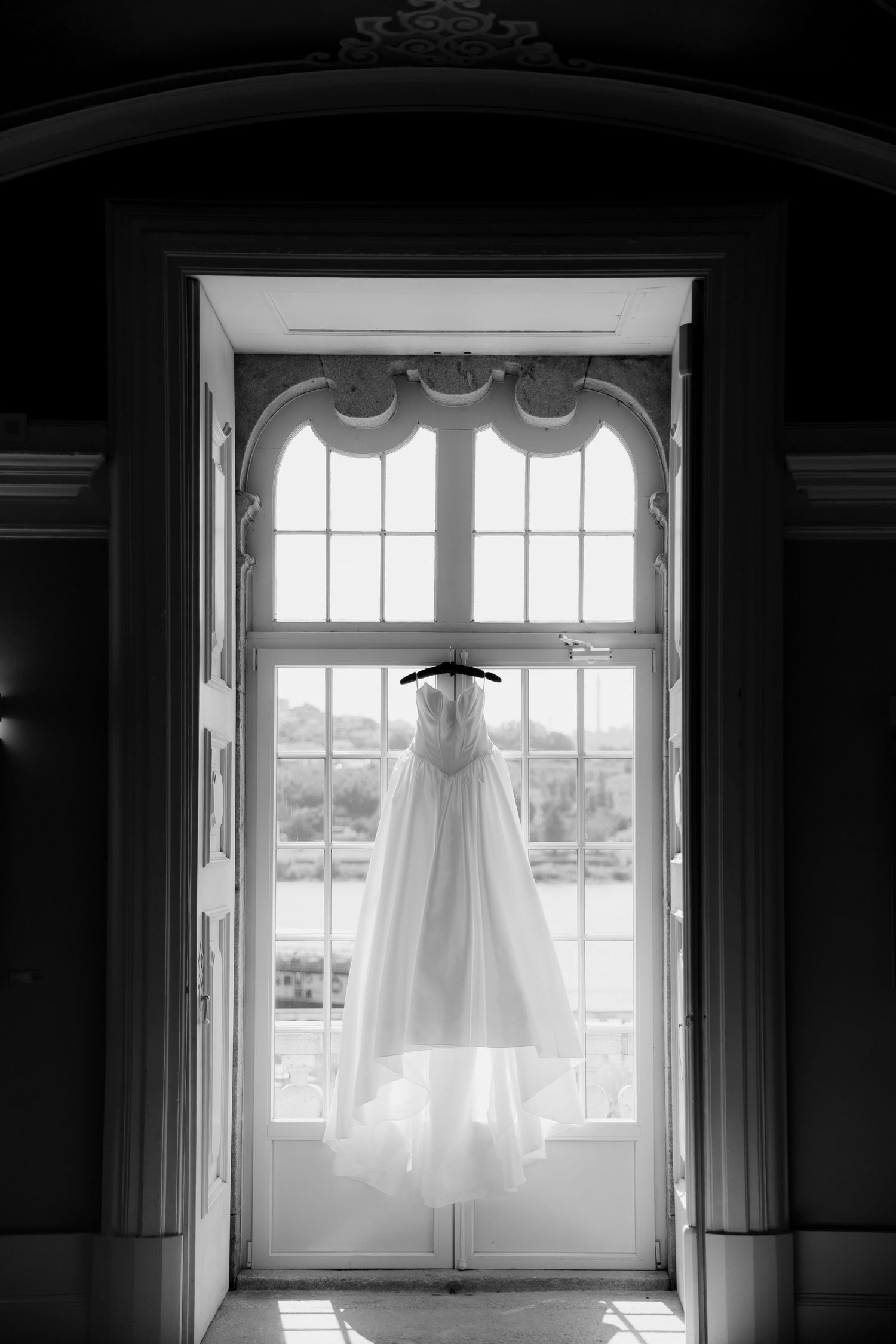 Black and white photo of a flowing white wedding dress hanging in an ornate arched window with natural light.
