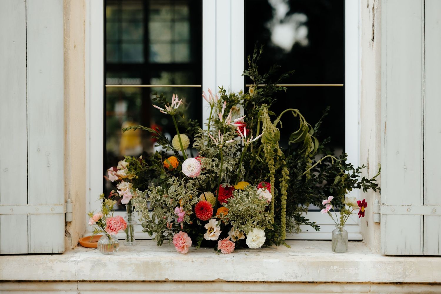 White shuttered windows decorated with floral arrangements and potted plants create a charming cottage exterior display.