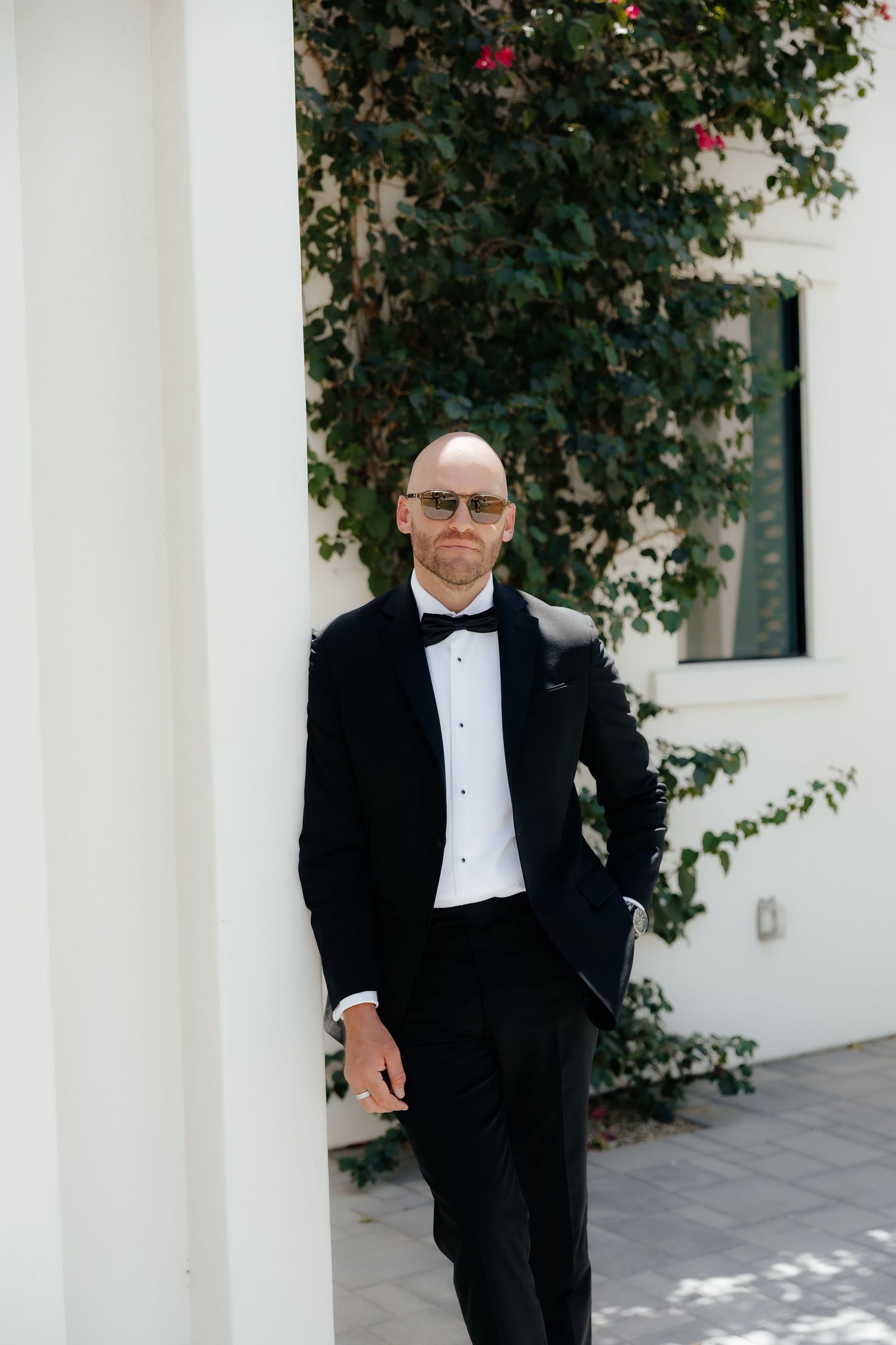 Bald groom in black tuxedo and bow tie leaning against white pillar with ivy backdrop.