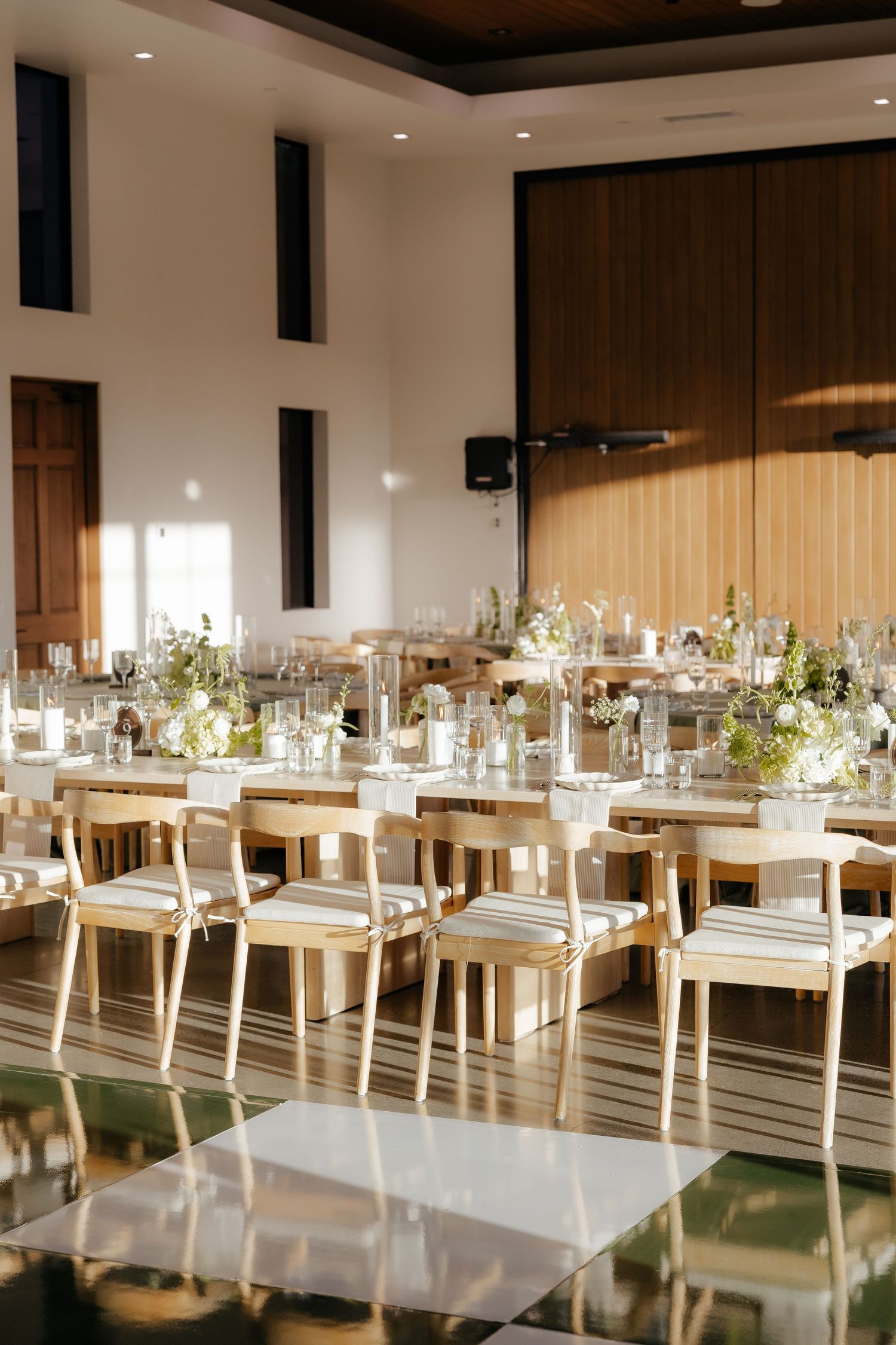 Bright modern wedding reception hall with long wooden tables, natural chairs, and green floral centerpieces.