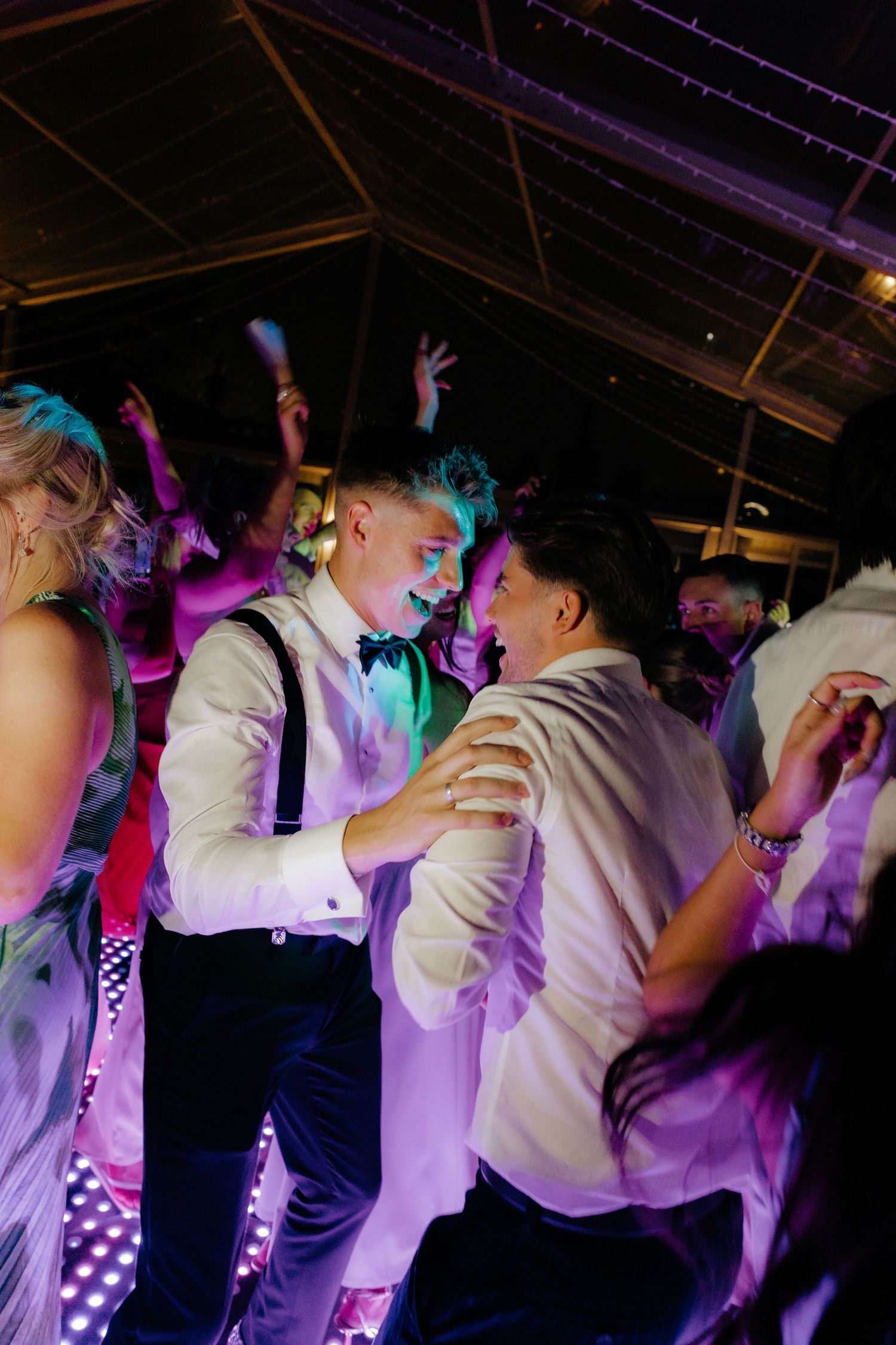 Wedding guests dance and celebrate on a dimly lit dance floor illuminated by purple and blue lighting.