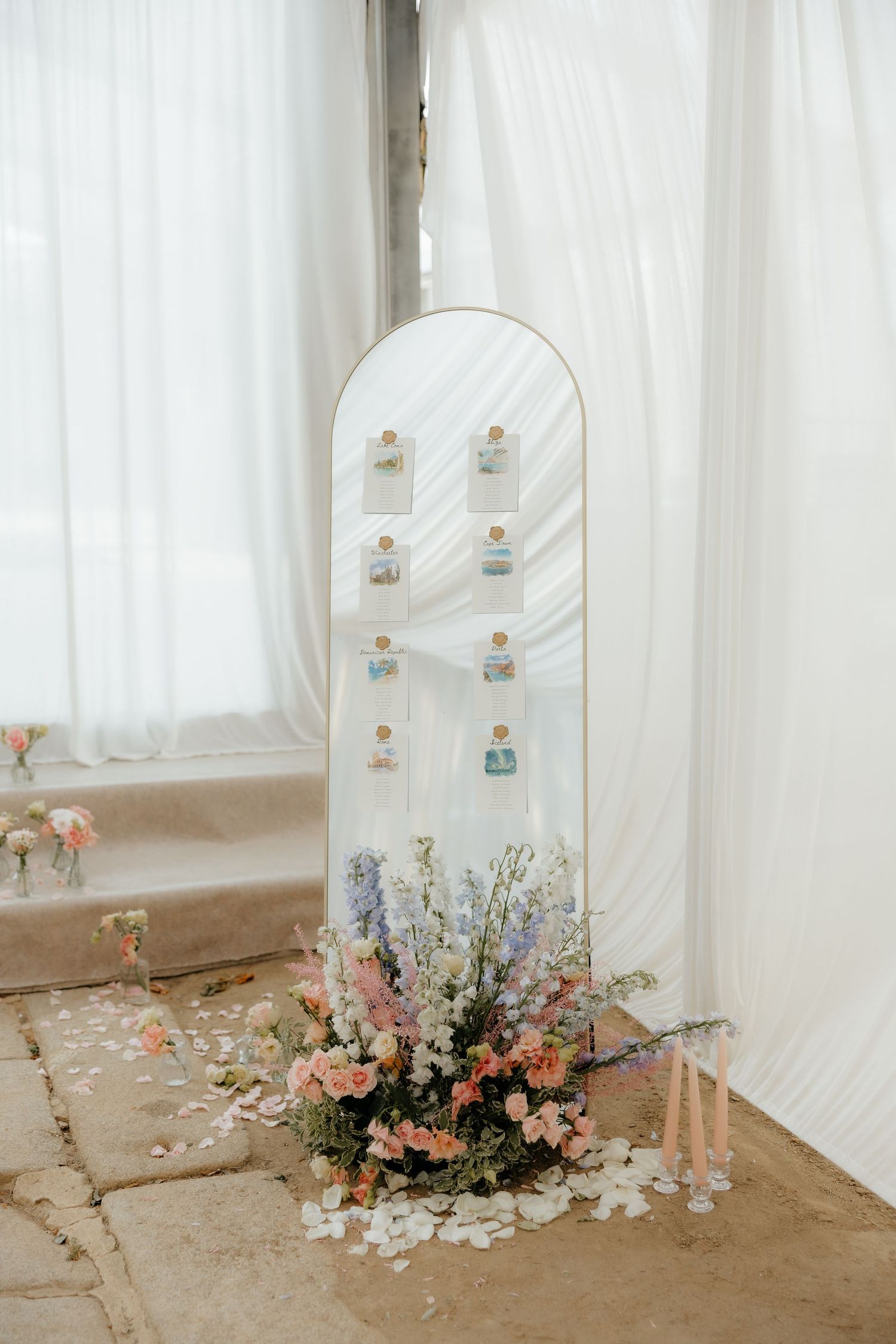 Elegant wedding seating chart display on an arch mirror with floral arrangement of pink and blue blooms at the base.