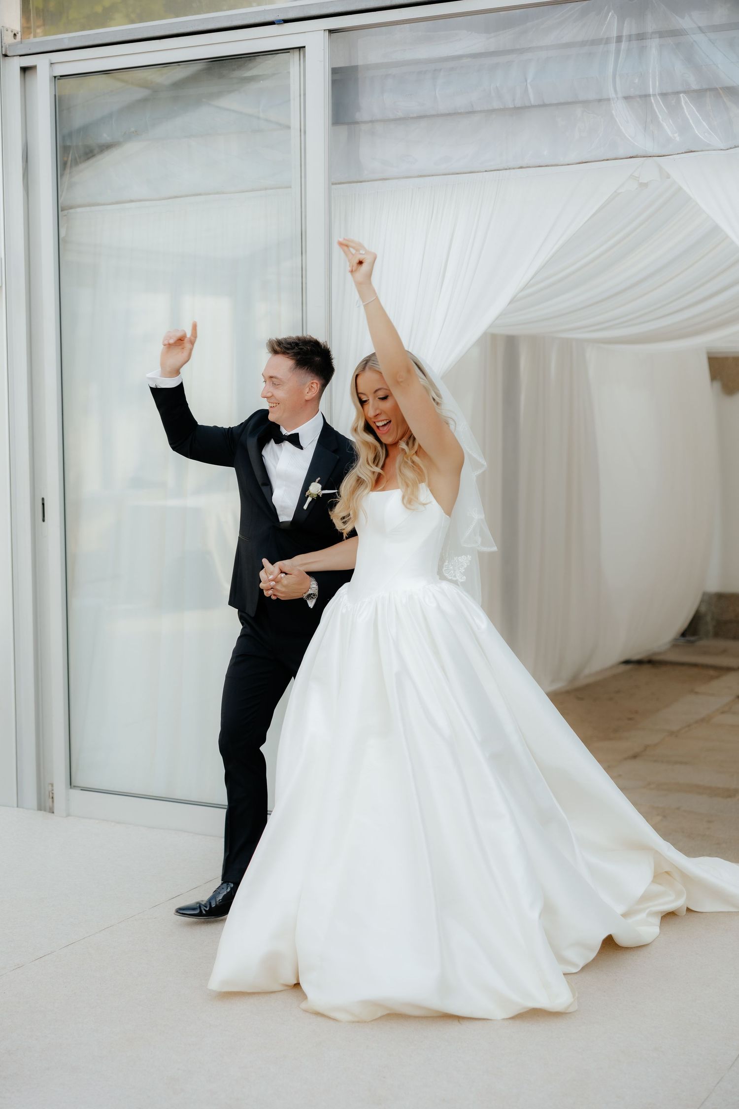 Newlywed couple dancing joyfully in a bright white room with flowing curtains and modern decor.