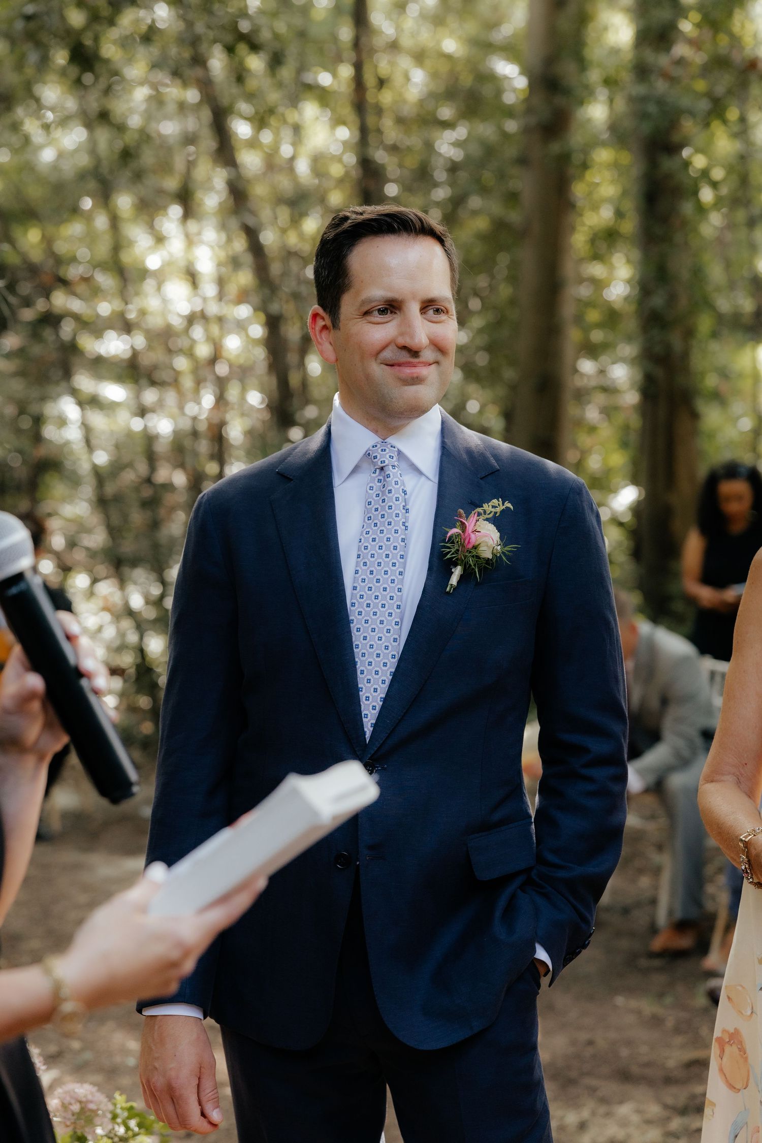 A groom in a navy suit and polka dot tie smiles during an outdoor wedding ceremony.