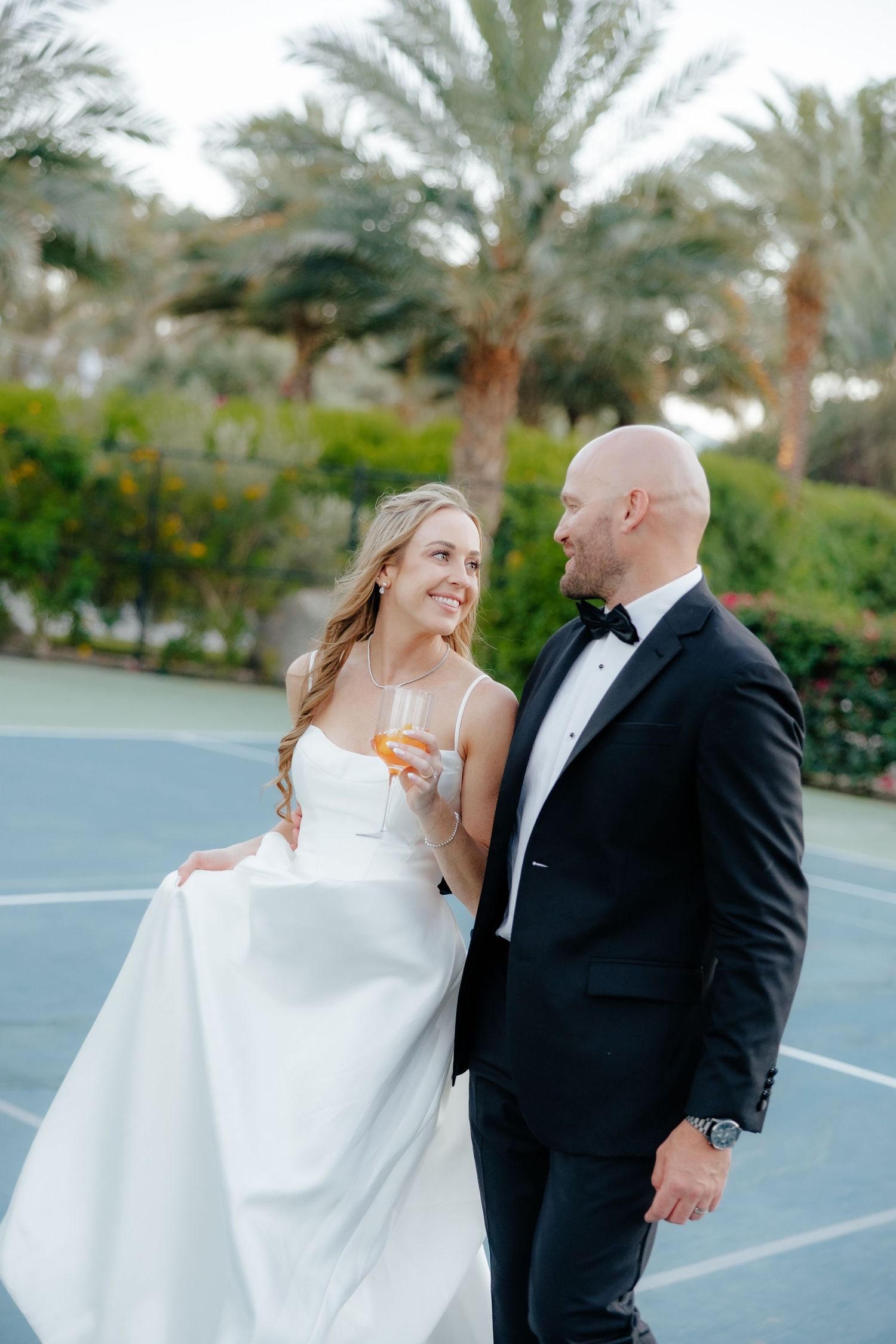 Bride and groom sharing romantic moment on tennis court, bride holding champagne glass among palm trees.