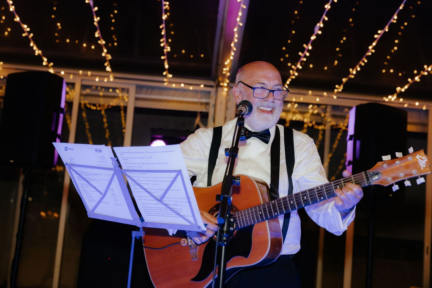 Musicians perform at an outdoor evening event under string lights with guitars and sheet music stands.