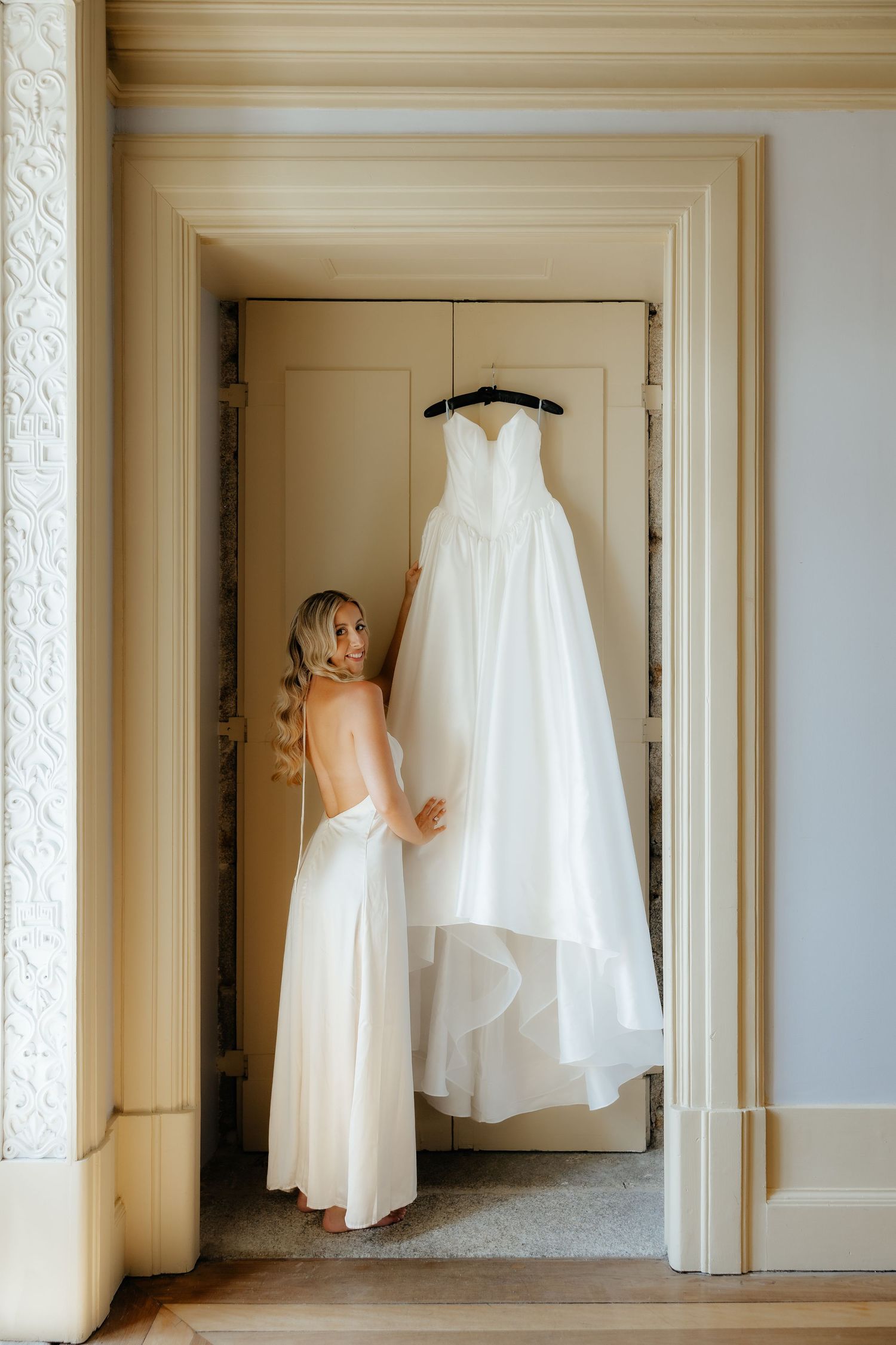 A series of elegant white wedding dresses displayed hanging in a bright, minimalist hallway with cream-colored walls.