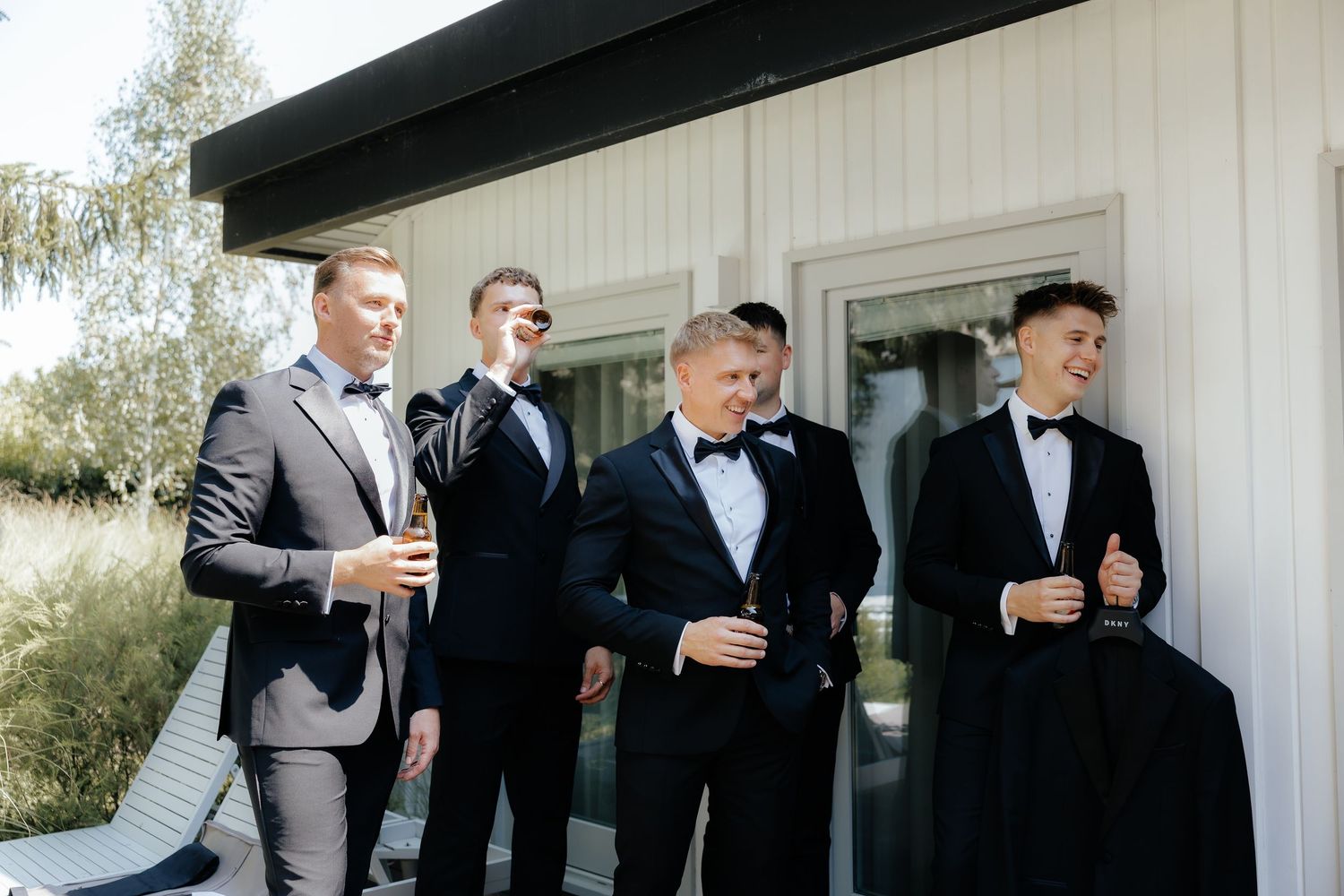 Groomsmen in black suits and tuxedos share drinks and laughter outside during a wedding celebration.