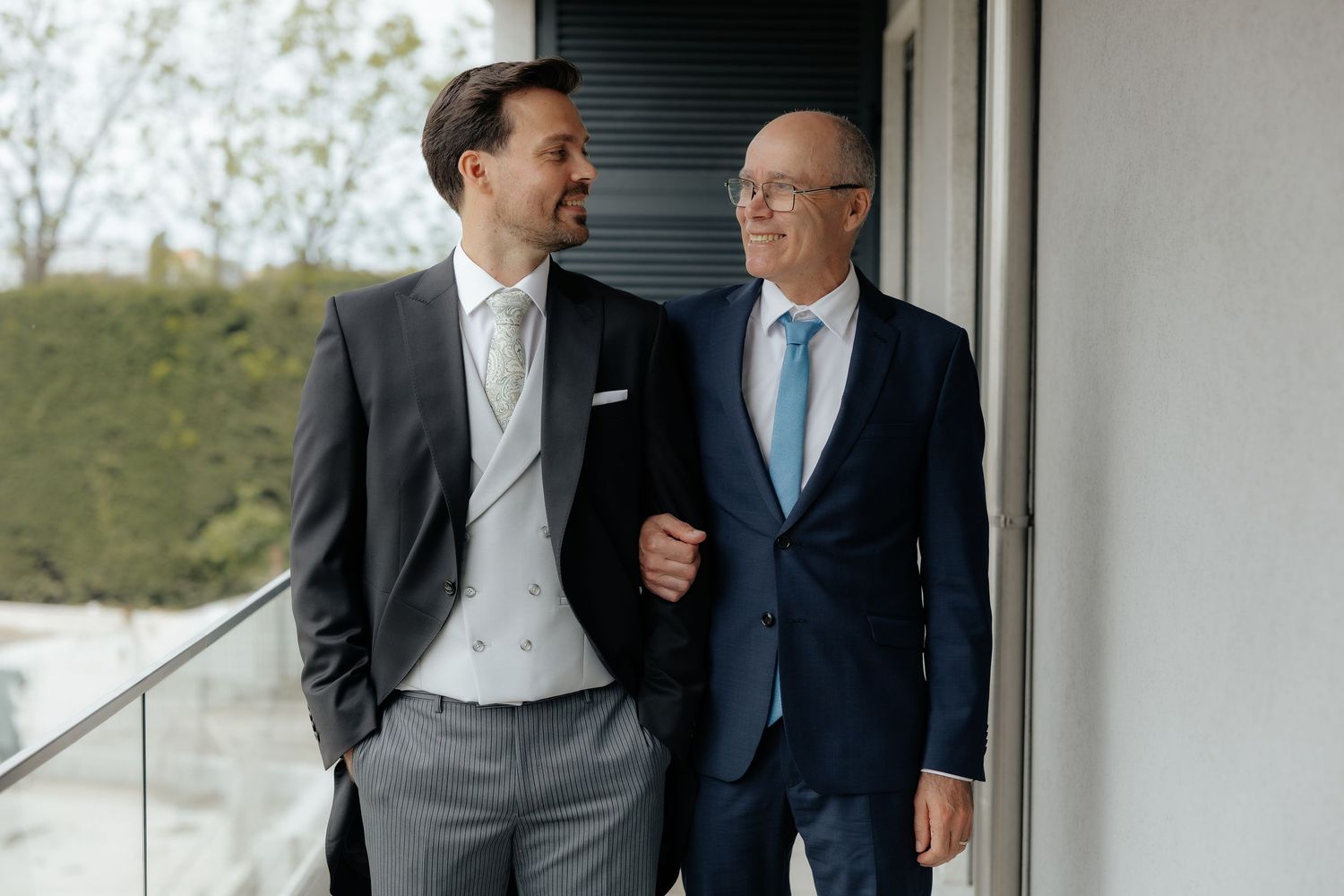 Groom and older man in navy suit share a smile on an outdoor balcony.
