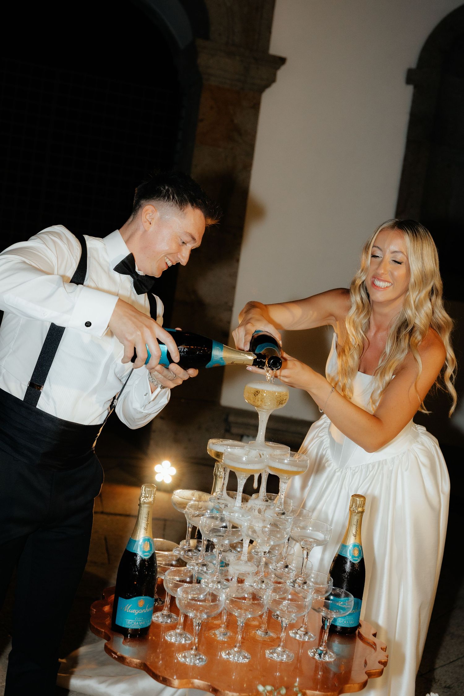 Bride and groom together pouring champagne into elegant tower of coupe glasses at wedding reception.
