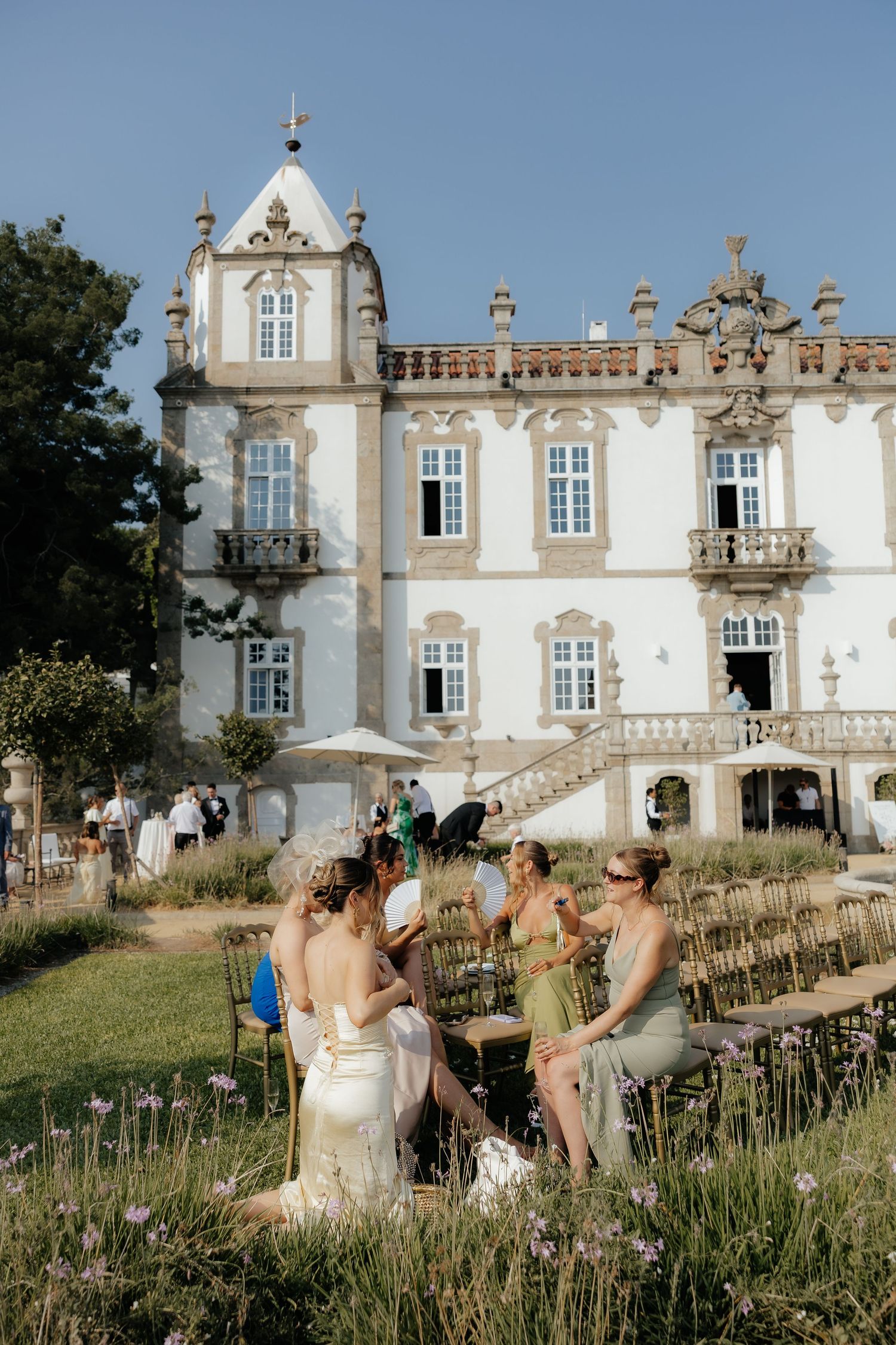 Bride and bridesmaids in sage dresses relax in garden before elegant white baroque wedding venue.