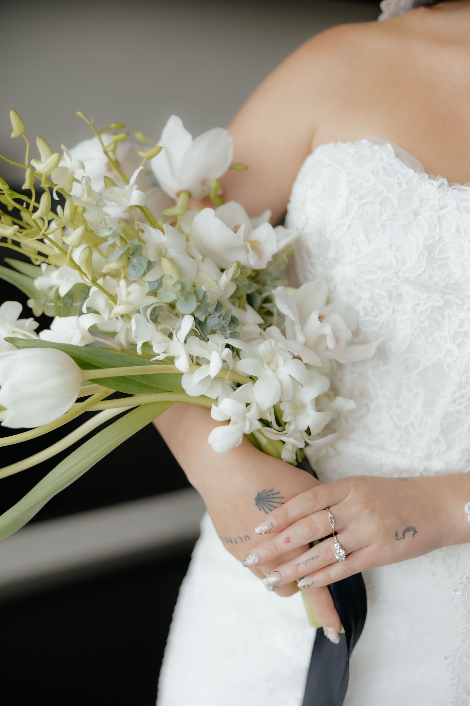 Bride in white lace gown holding elegant bouquet of white tulips and orchids, showcasing diamond engagement ring and delicate tattoos.