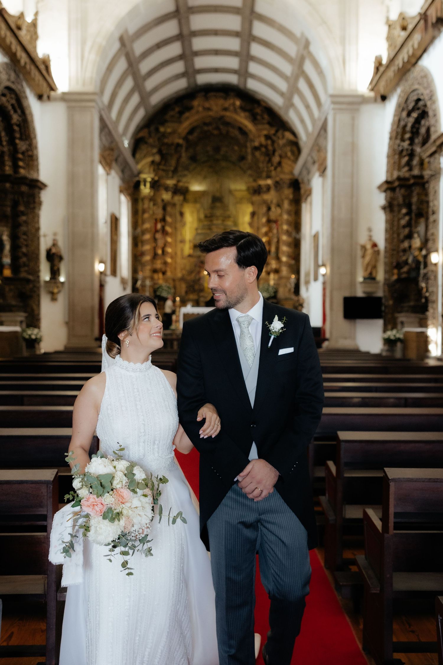 Newlyweds walk down church aisle smiling at each other, bride holding a bouquet.