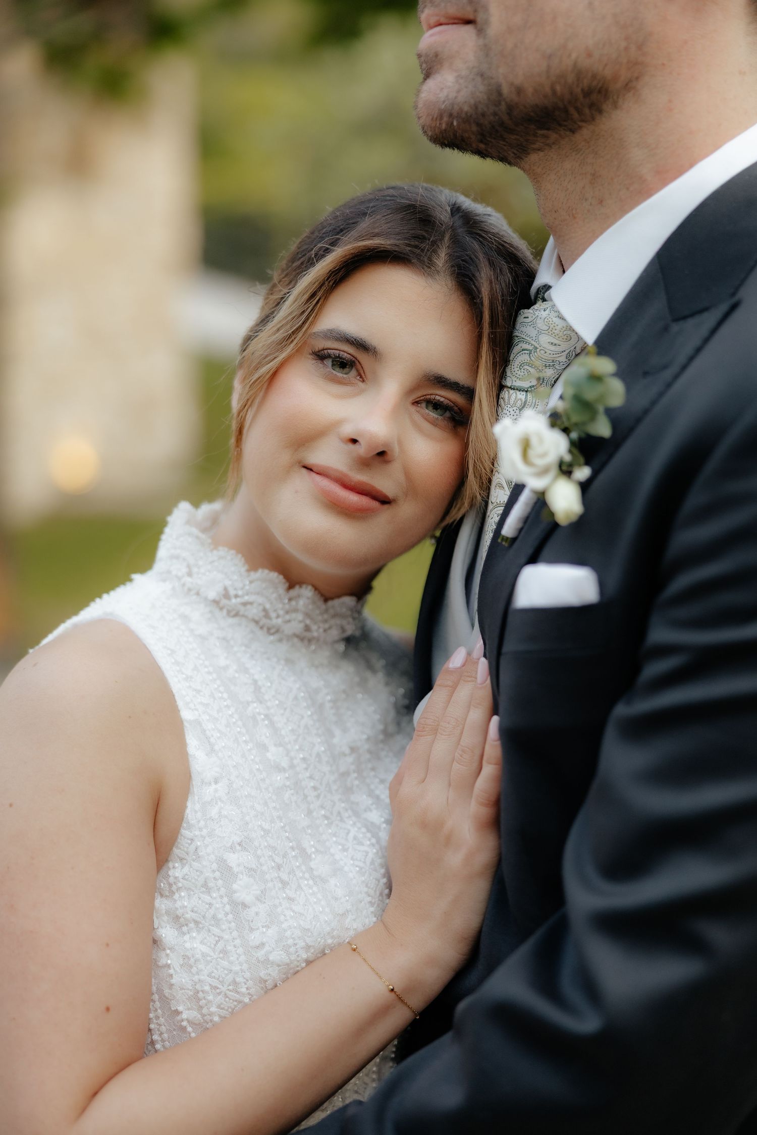 Bride in white lace dress leans against groom in black suit with floral boutonniere at outdoor wedding.