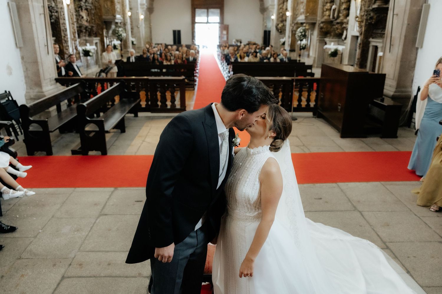 Newlyweds share first kiss at church altar with guests seated along red carpet aisle.