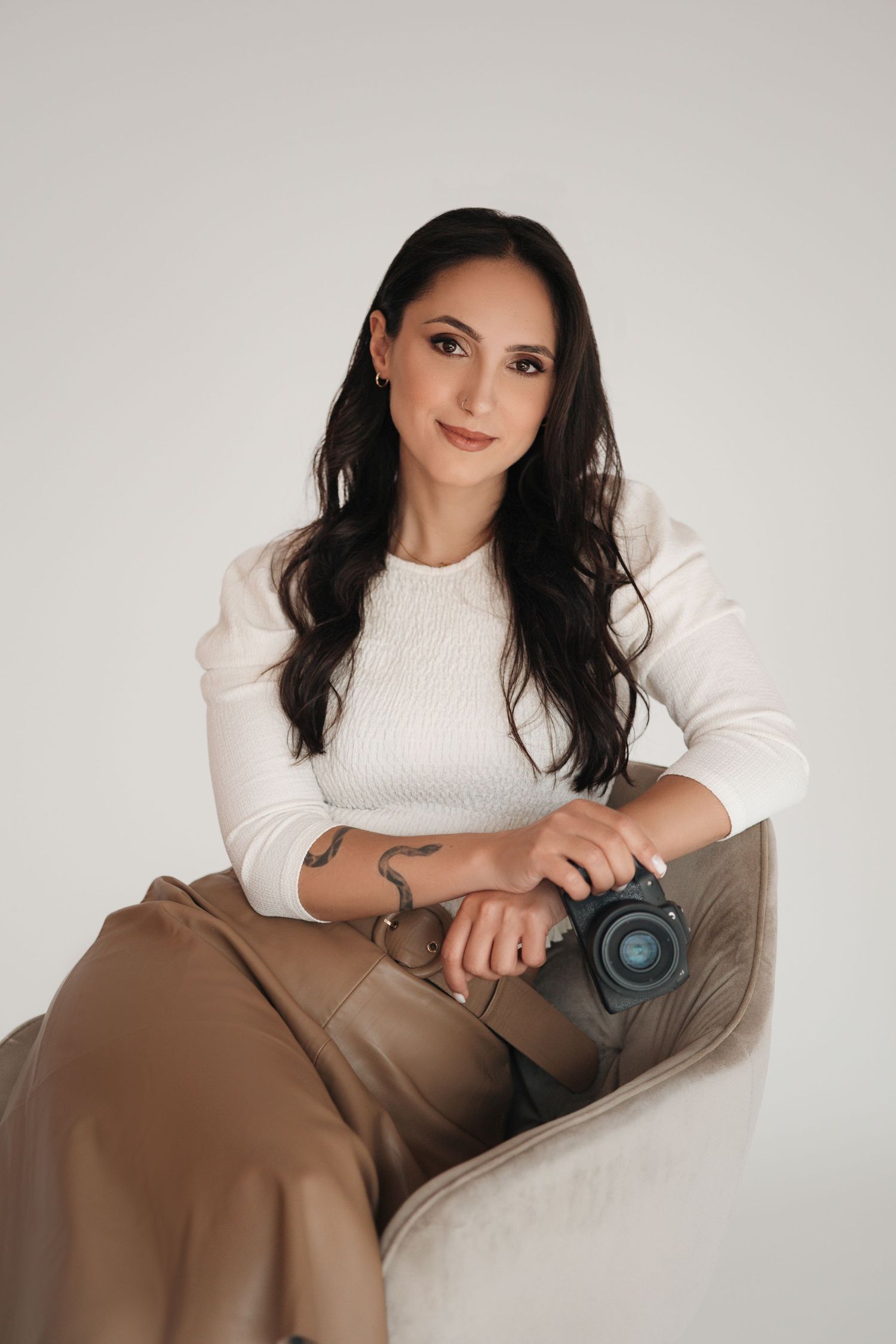 Professional female photographer sitting in chair holding camera wearing white sweater and tan pants.