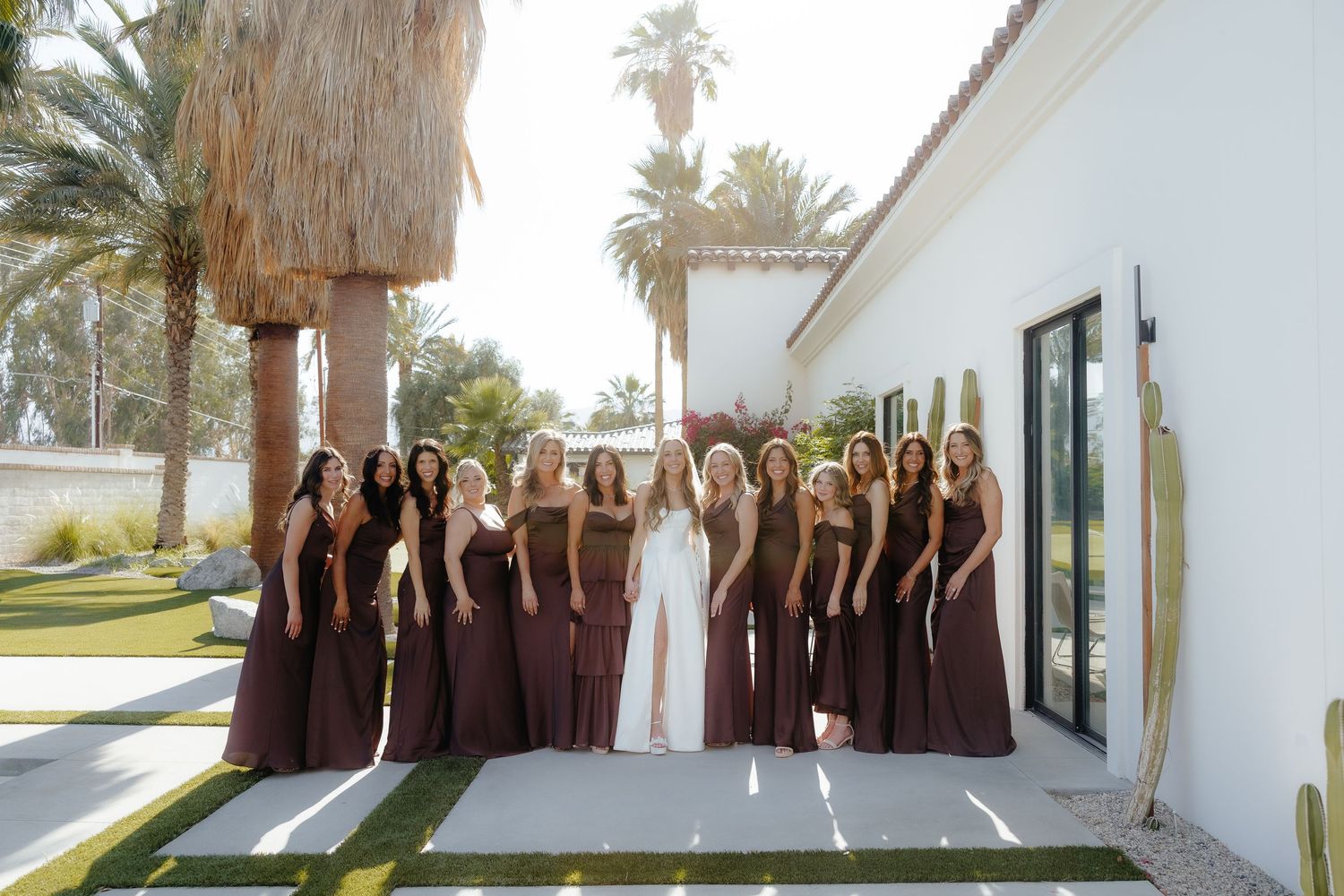 Bride surrounded by bridesmaids in dark brown gowns posing outside a white building.