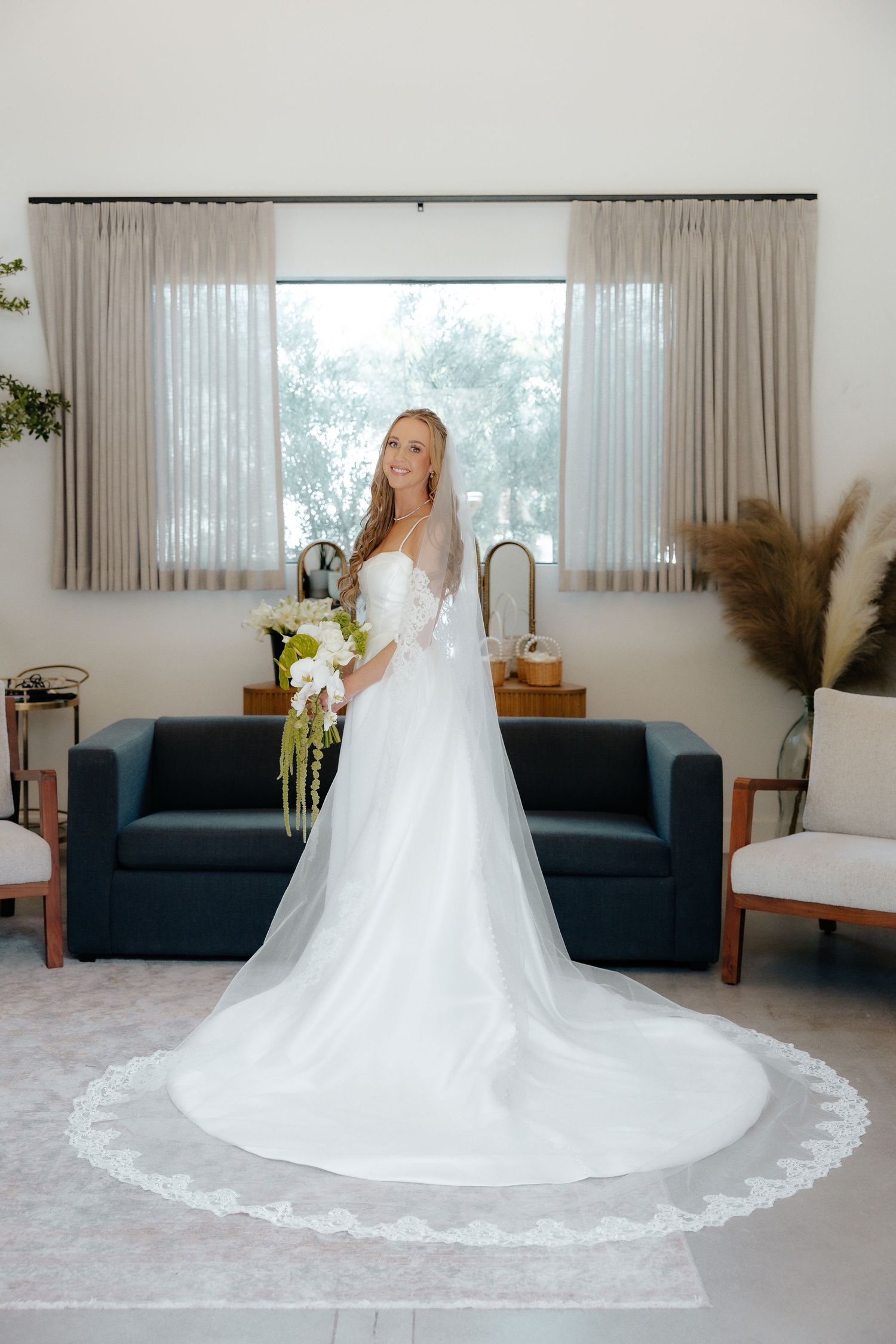 Bride in lace-trimmed wedding gown with long veil posing in a sunlit bridal suite.